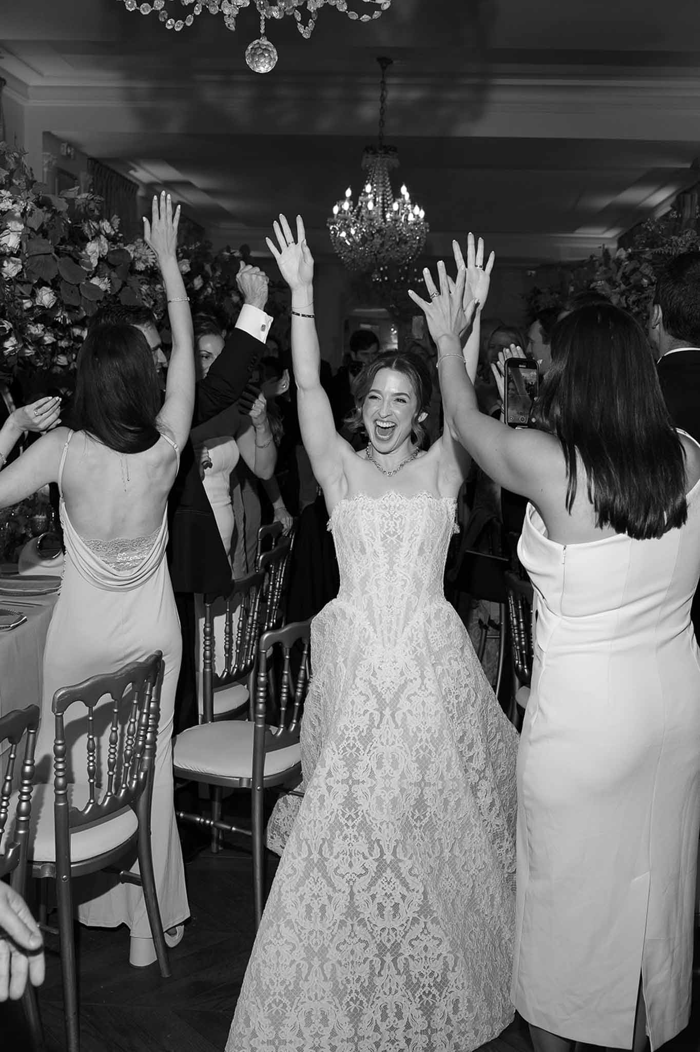Black and white image of bride dancing with arms raised surrounded by guests at indoor reception