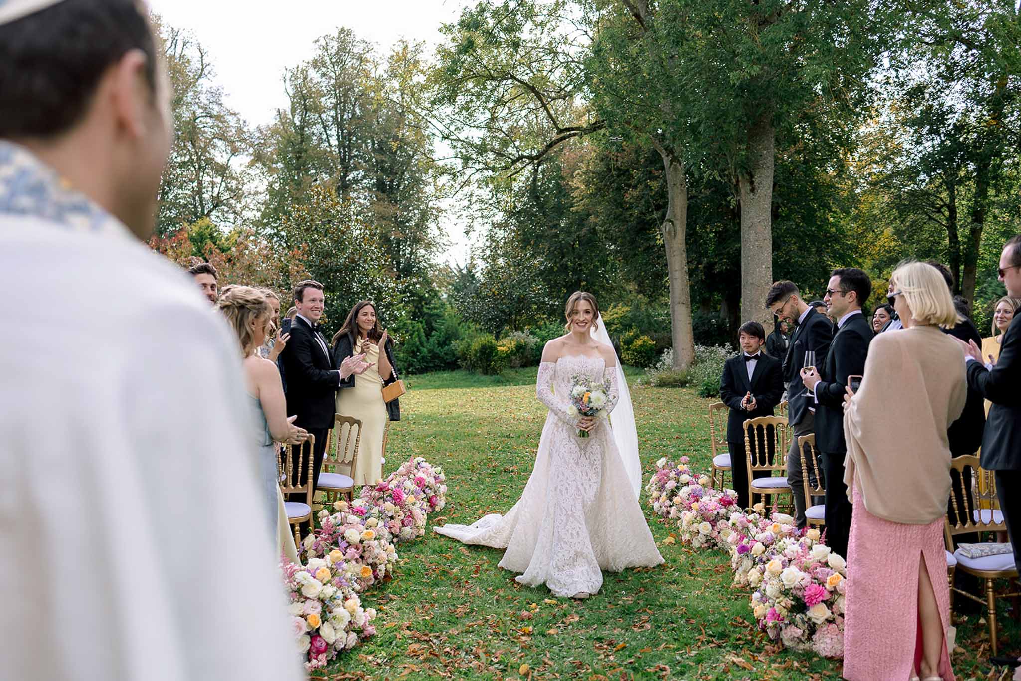Bride walks down aisle in lace gown past hot pink and blush floral ground arrangements with standing guests