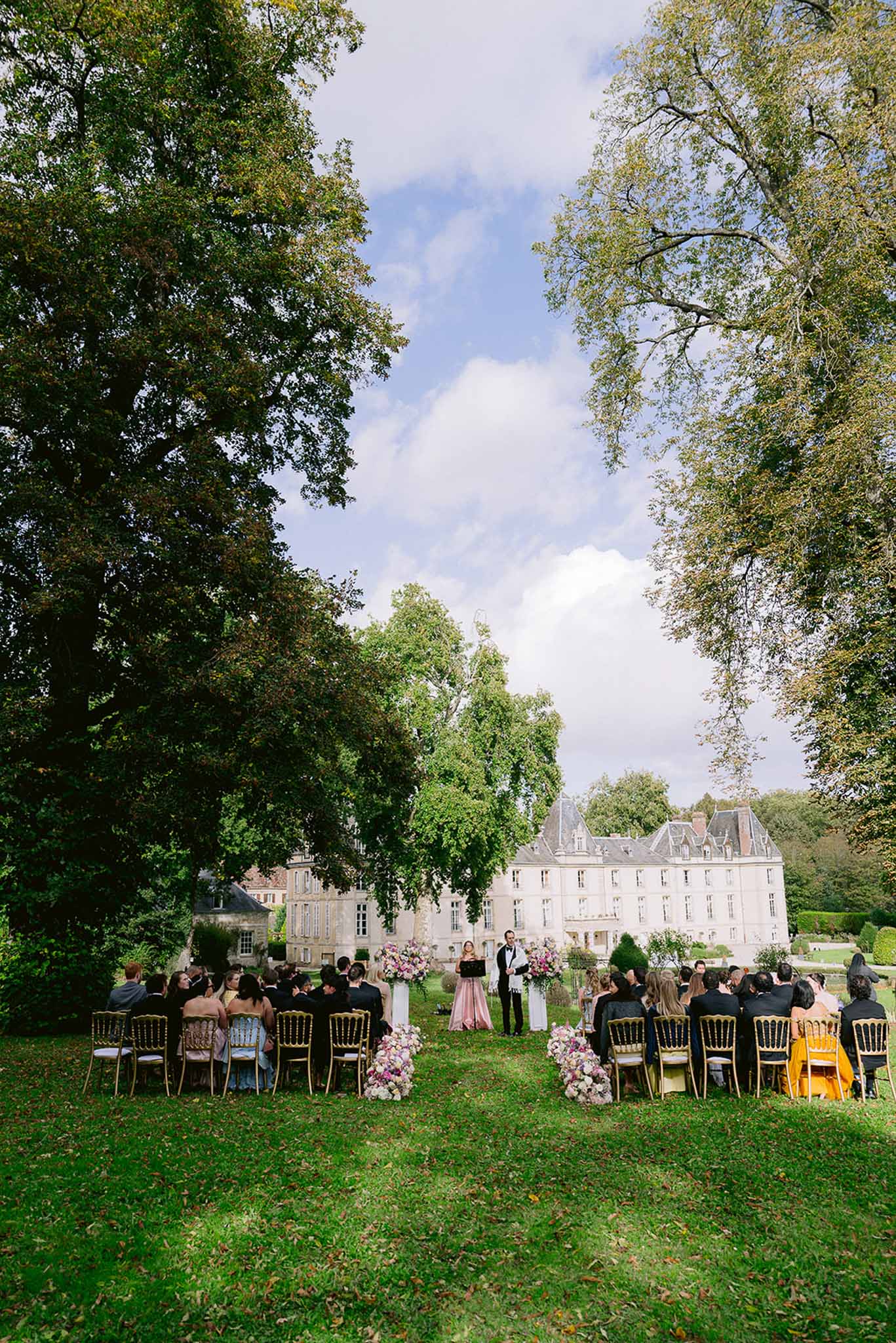Outdoor wedding ceremony at a French chateau with guests seated in gold Chiavari chairs and pink and white floral aisle decor