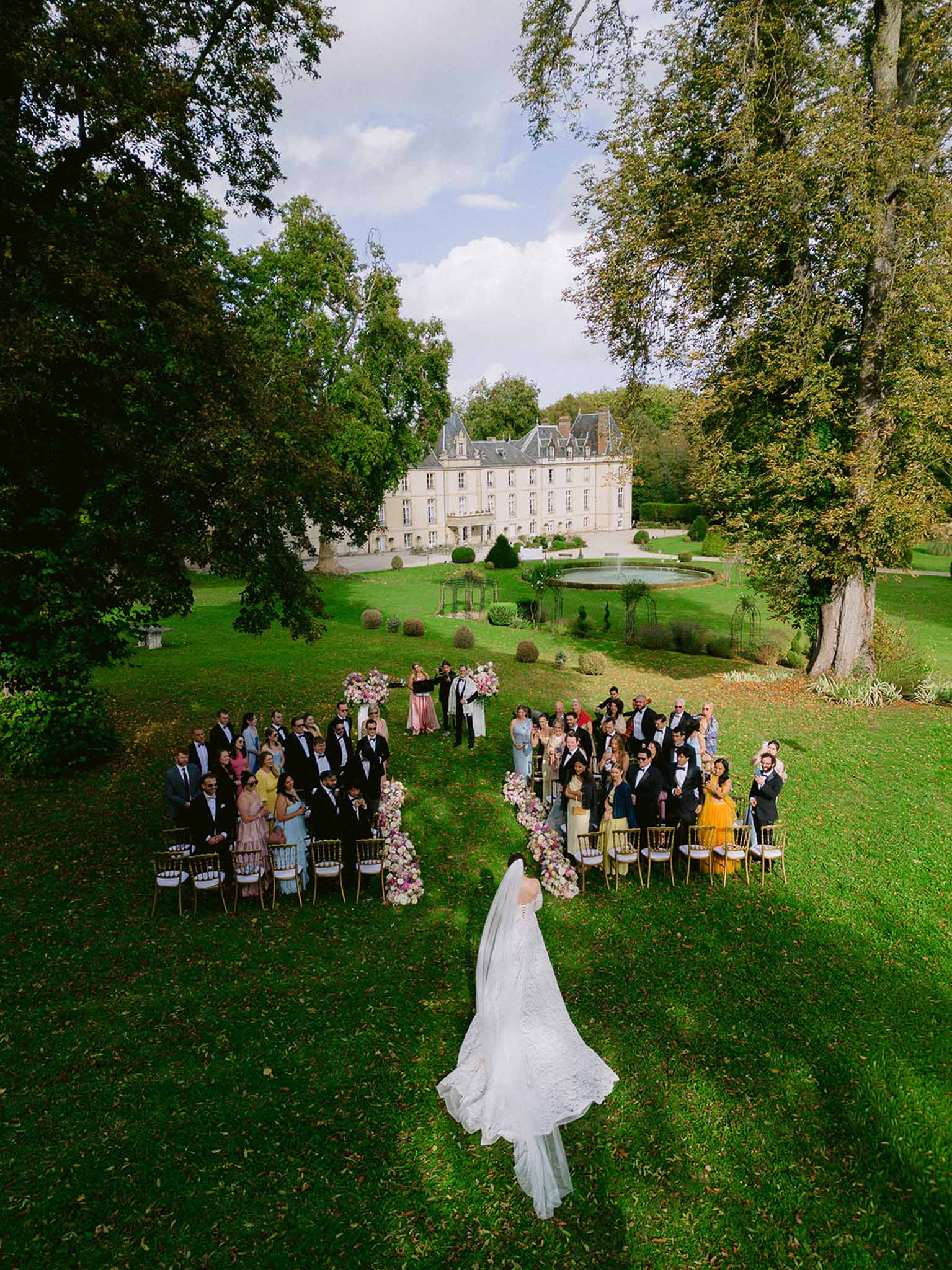 Aerial view of bride walking aisle past 50 guests with pink floral stands before classical chateau