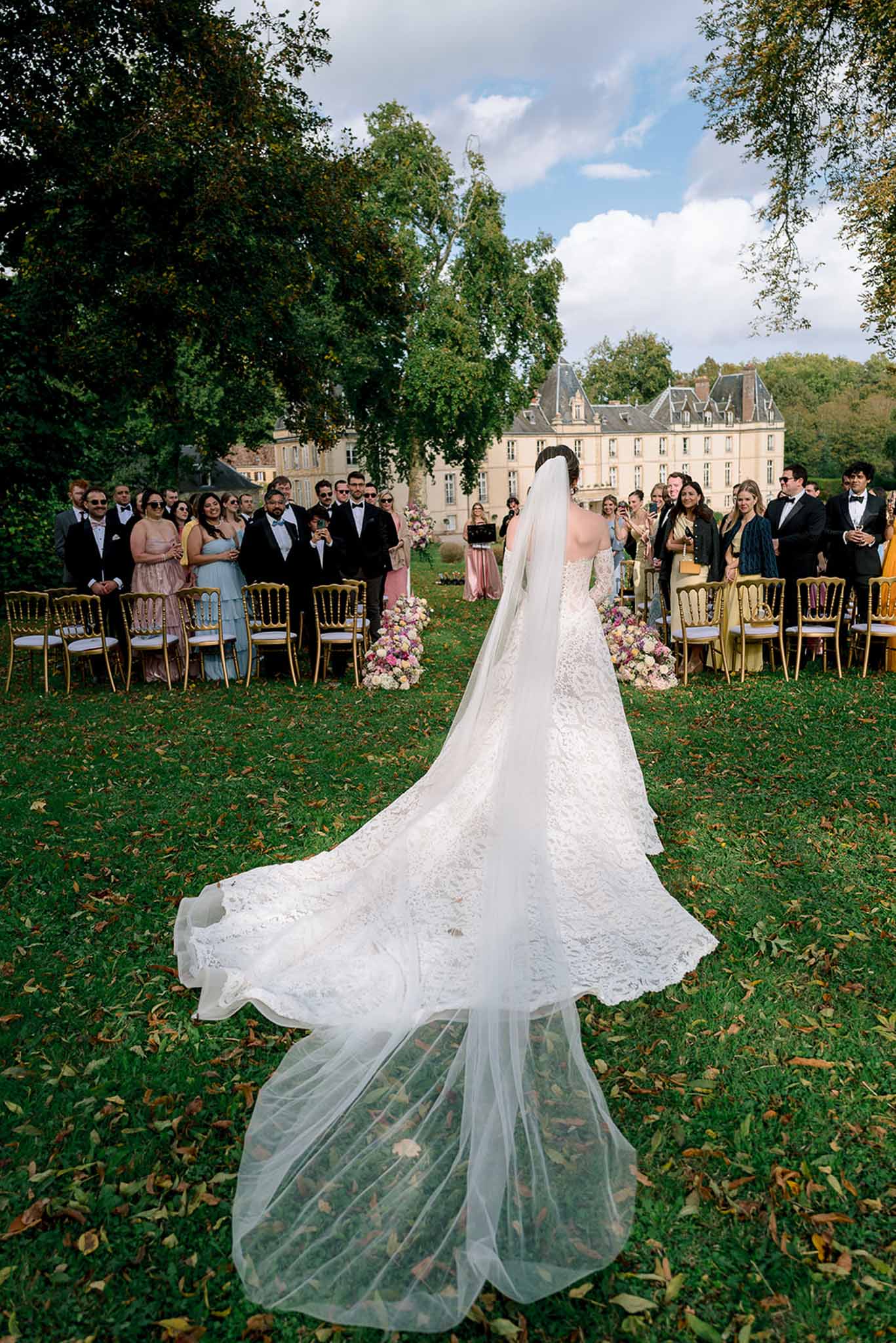 Bride walking down aisle from behind with cathedral train and veil, pink floral markers and chateau beyond