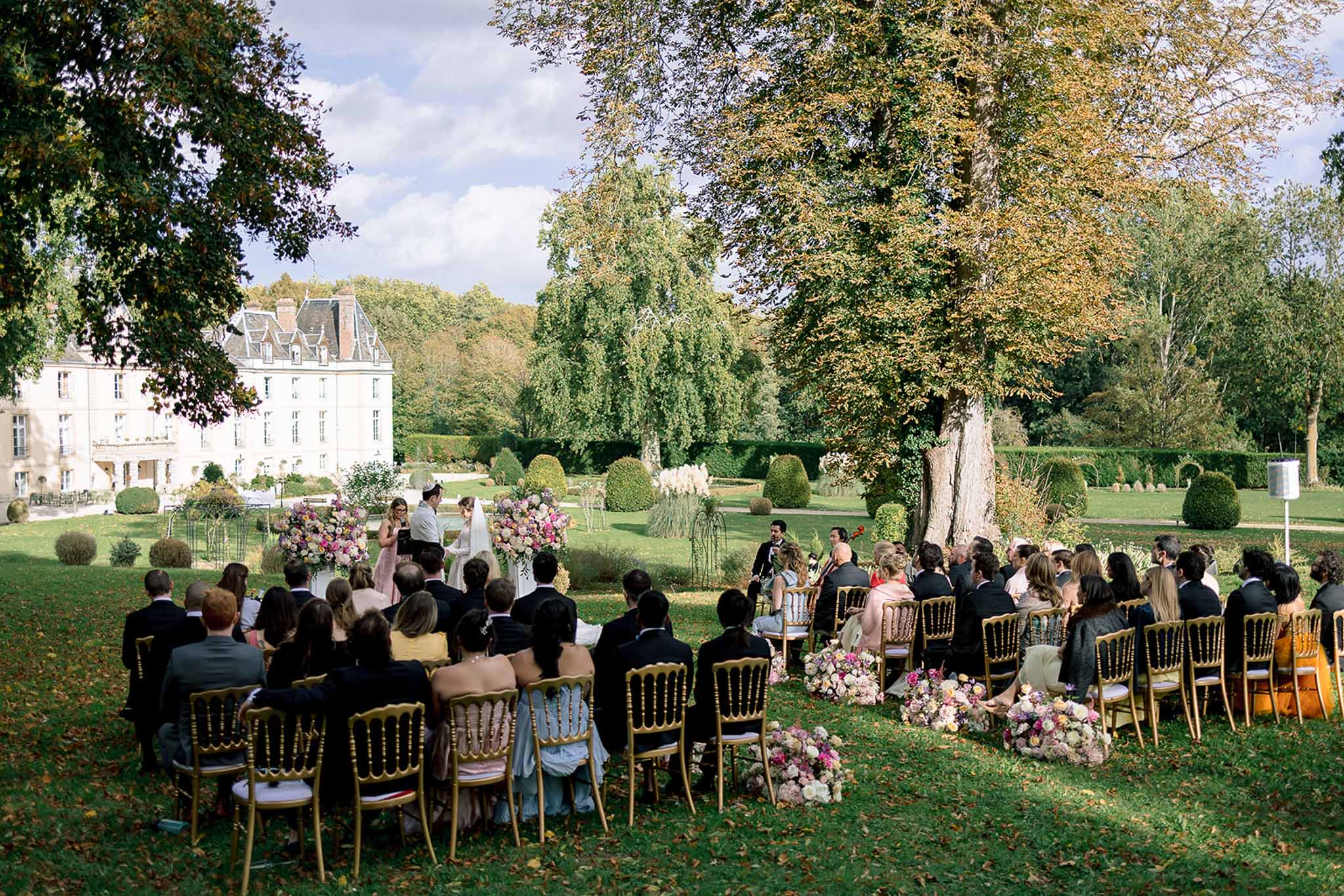 Sixty guests at gold chiavari chairs watching vow exchange with pink floral altar before chateau