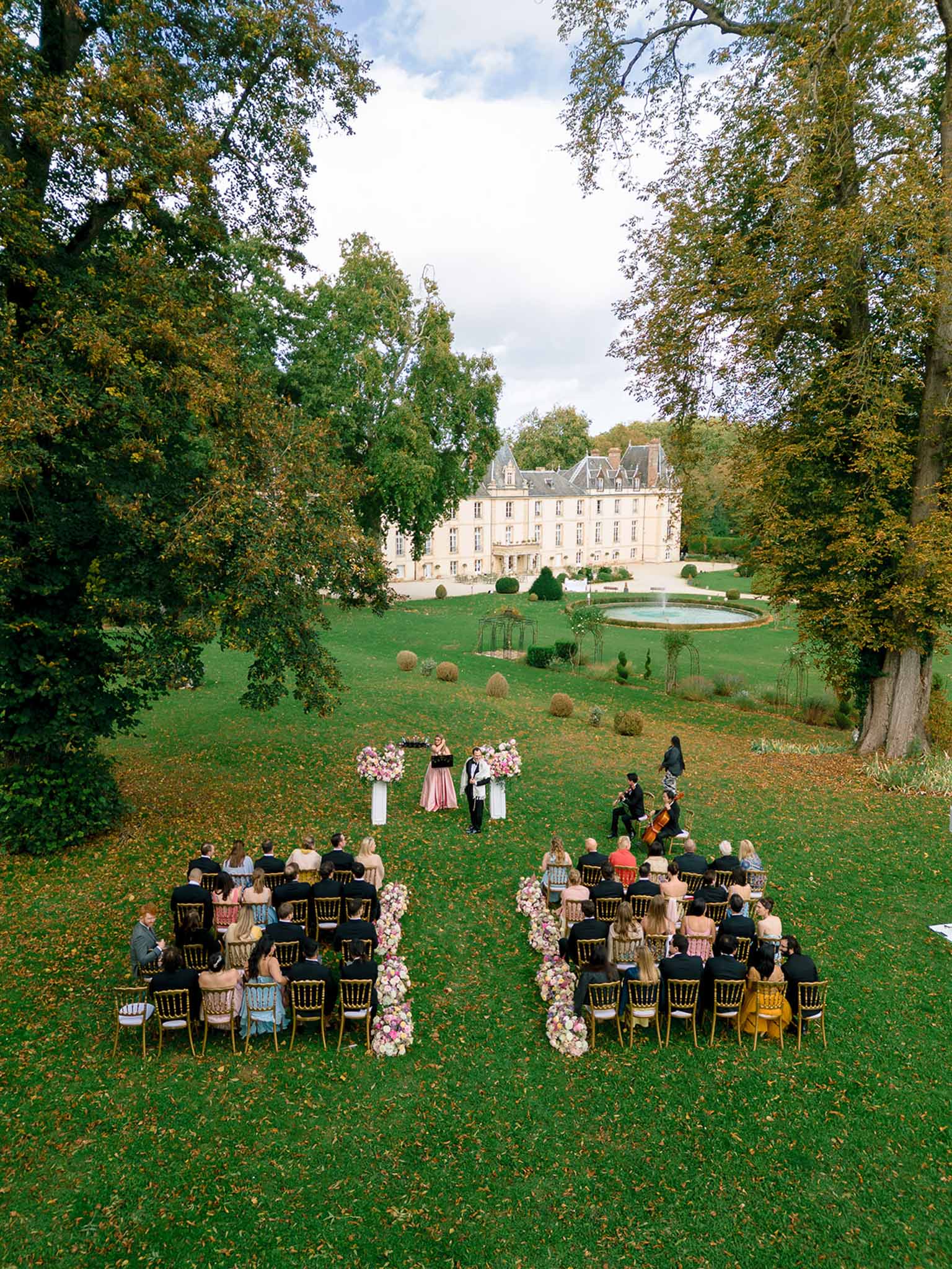 Aerial ceremony with pink floral column arrangements, 50 guests on gold chairs, and chateau fountain garden