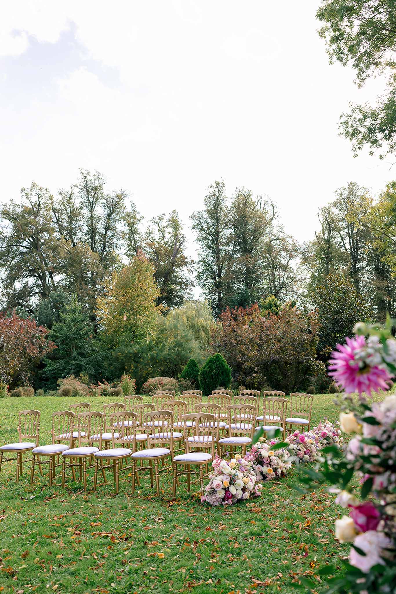Curved rows of gold Napoleon chairs with ground-level blush and burgundy rose arrangements lining garden ceremony aisle