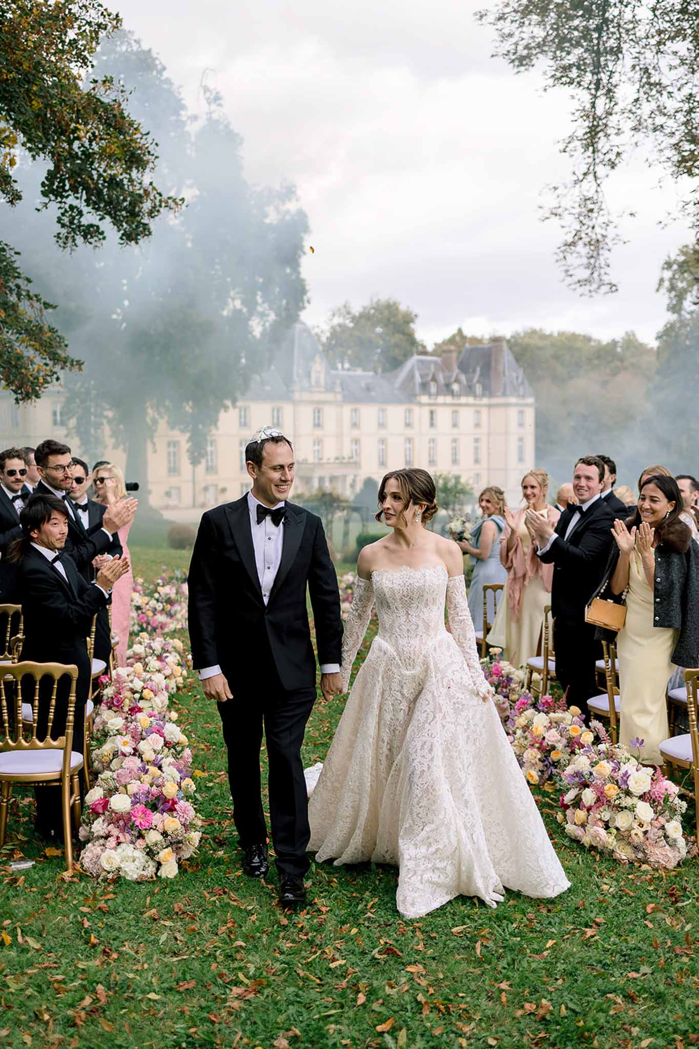 Couple walks back up aisle through smoke with multicolored floral borders and guests on gold chairs