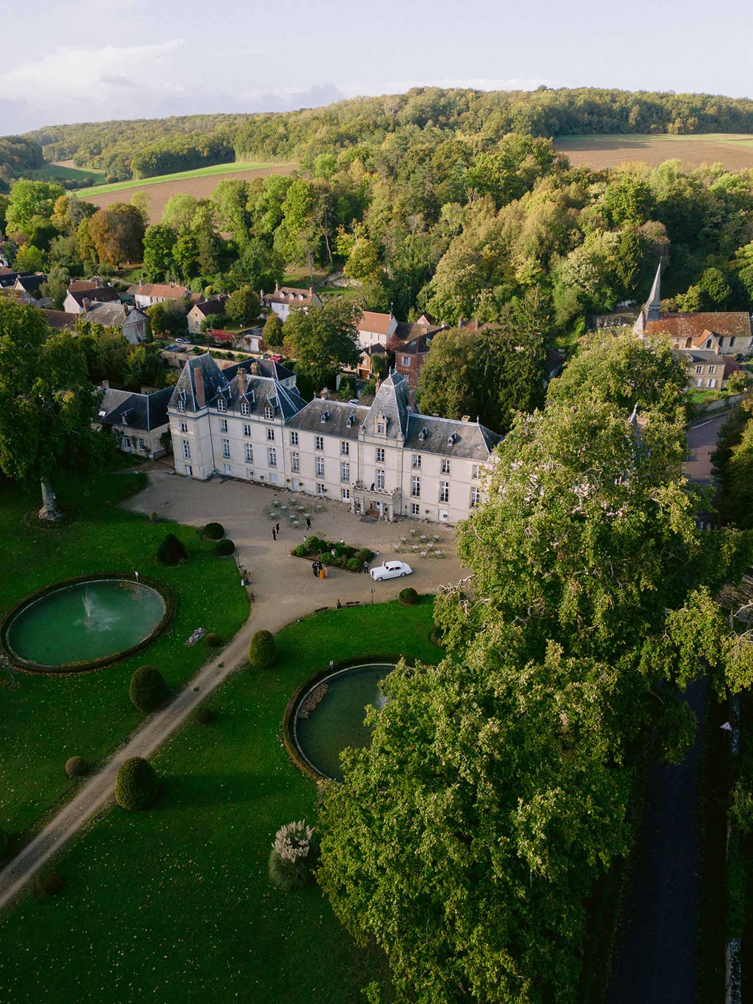 Aerial view of cream limestone chateau with formal gardens, twin ponds, and village beyond