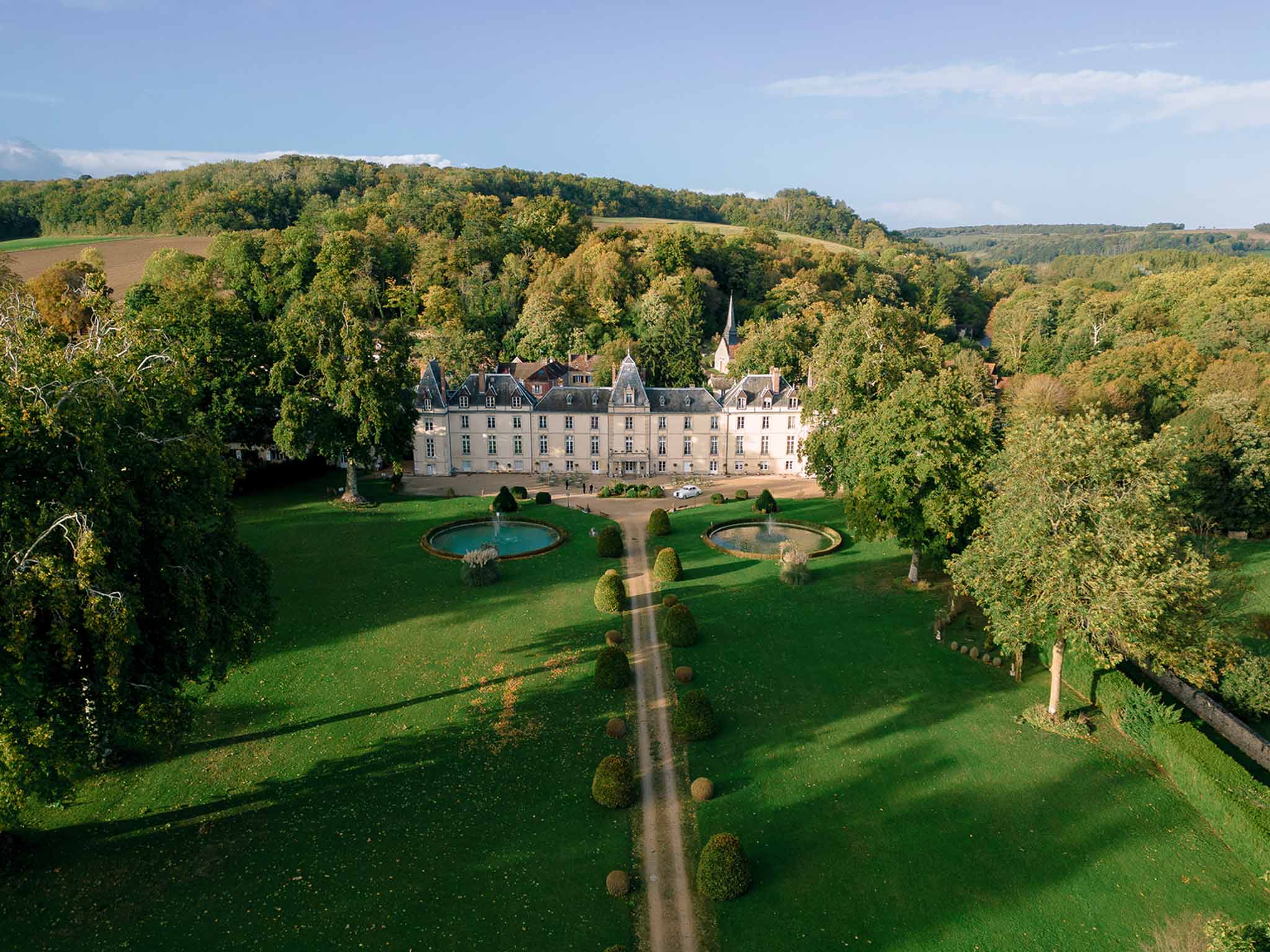 Aerial view of classical chateau with turrets, topiary-lined driveway, twin fountains, and chapel spire