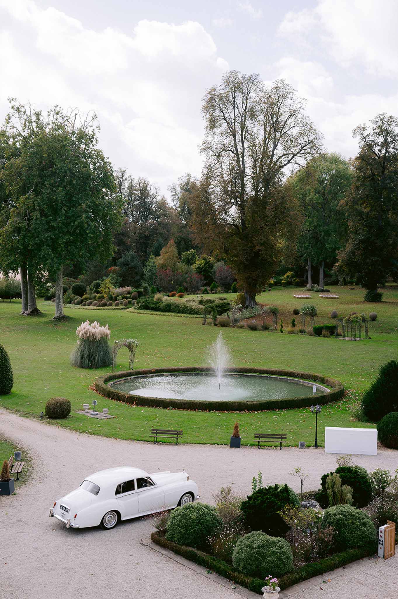 Chateau garden with circular fountain, vintage white Rolls-Royce on gravel, clipped hedges, and autumn parkland