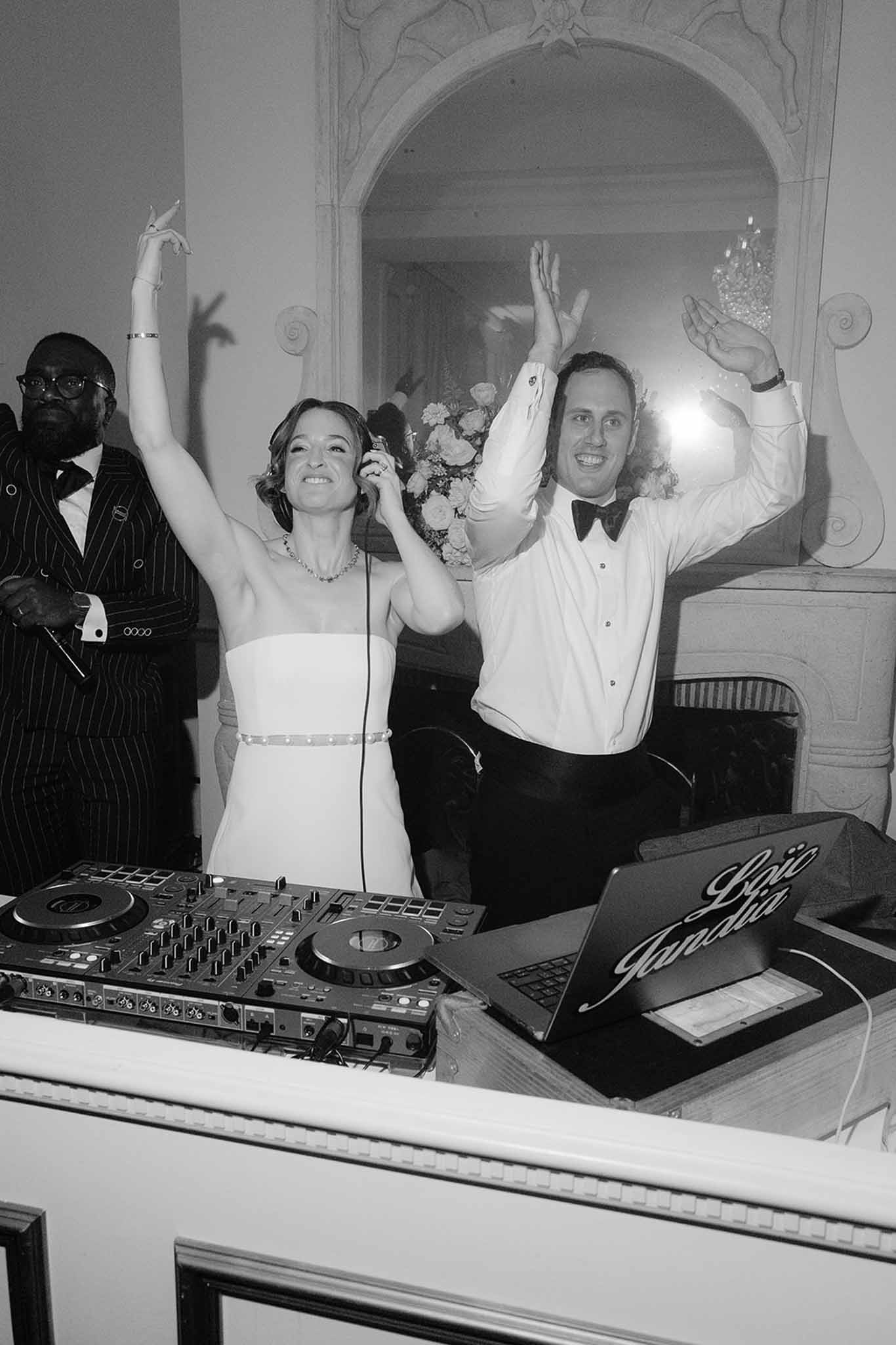 Black and white shot of bride and groom behind DJ booth with arms raised in chateau ballroom