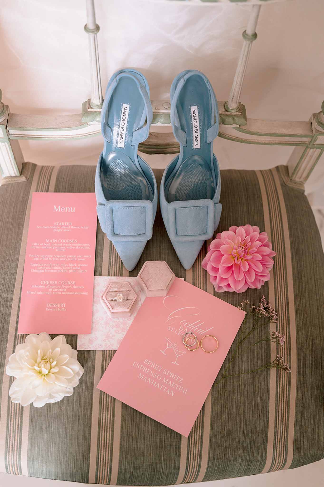 Blue Manolo Blahnik heels with coral menu cards, blush ring box, gold bands, and pink dahlia