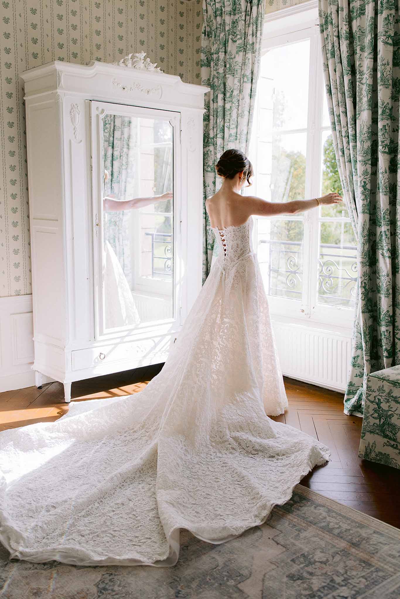 Bride in lace gown with cathedral train opening curtains at tall window in toile de Jouy room