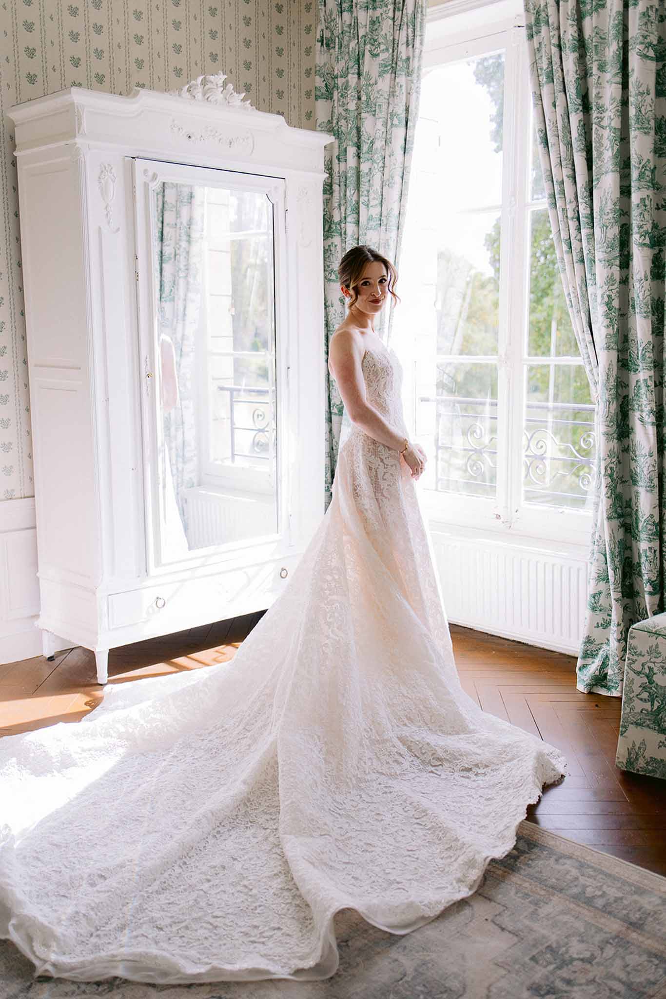 Bride in strapless ivory lace gown with cathedral train looking over shoulder in toile de Jouy chateau room