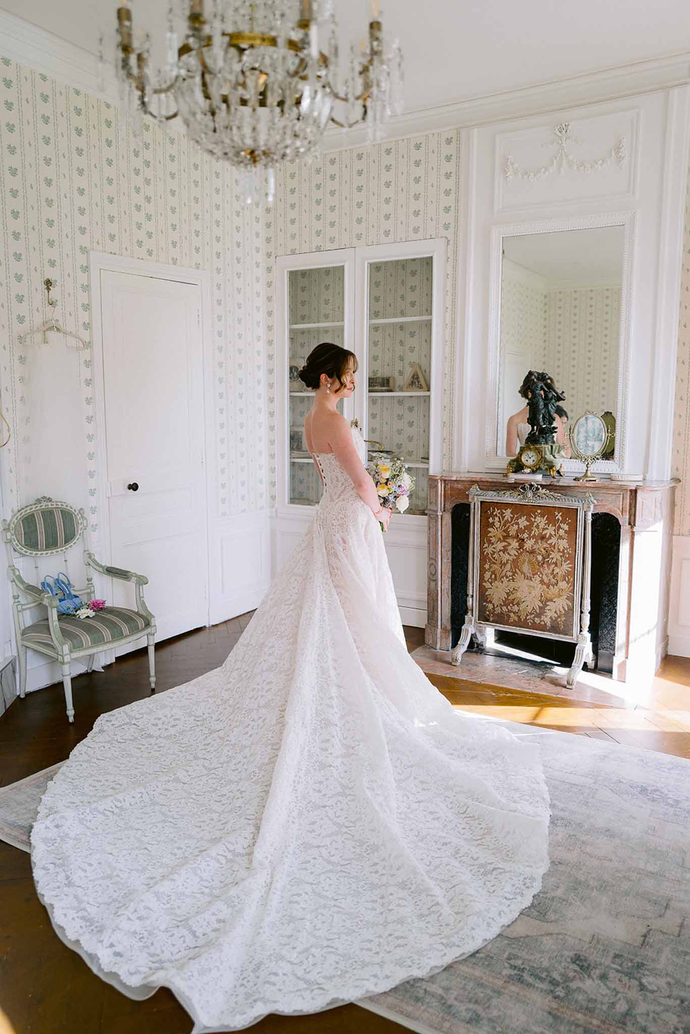 Bride in strapless ivory lace gown with cathedral train standing in chateau room with crystal chandelier and marble fireplace