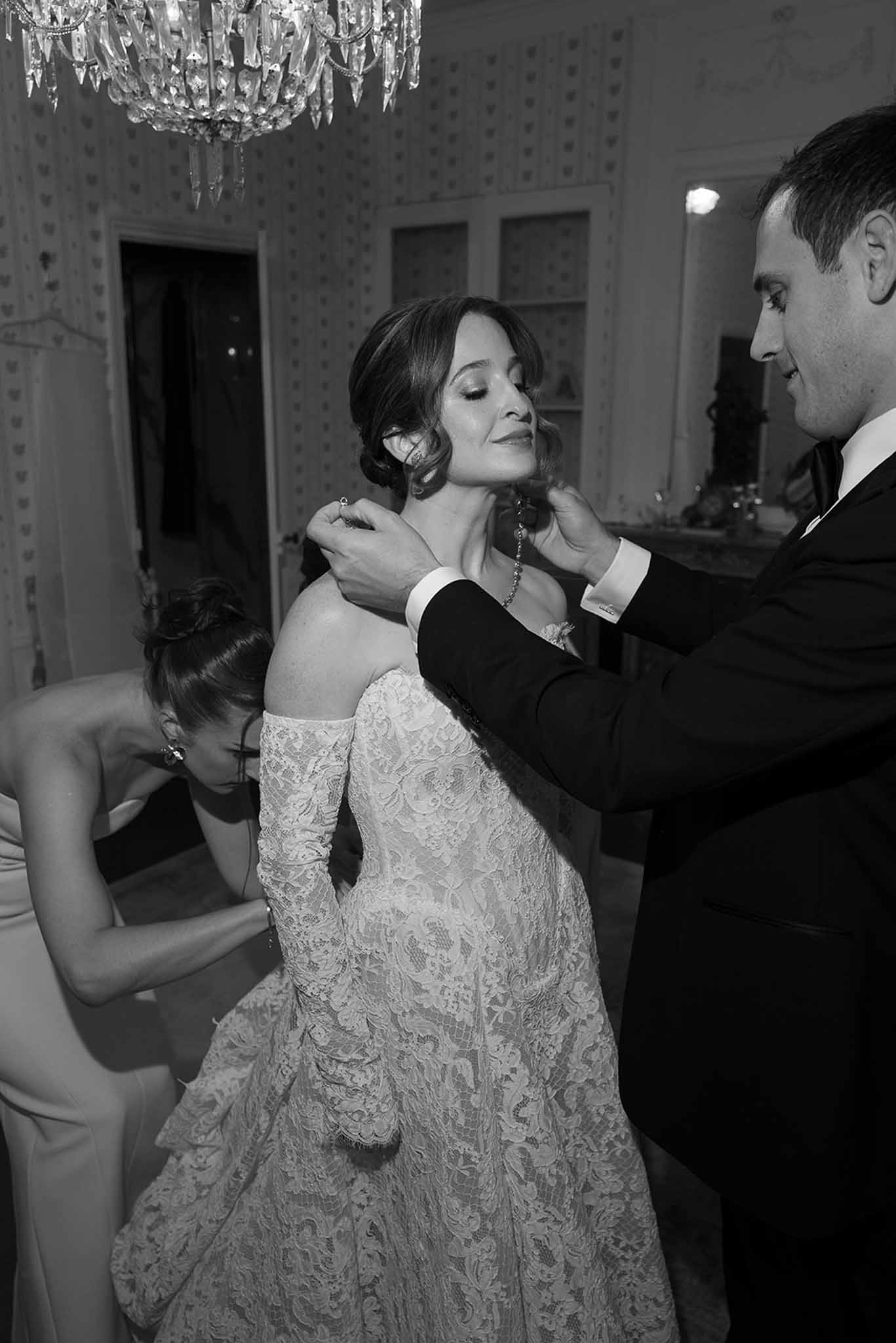 Black and white of man fastening bride's earring as woman adjusts gown in wallpapered room with crystal chandelier