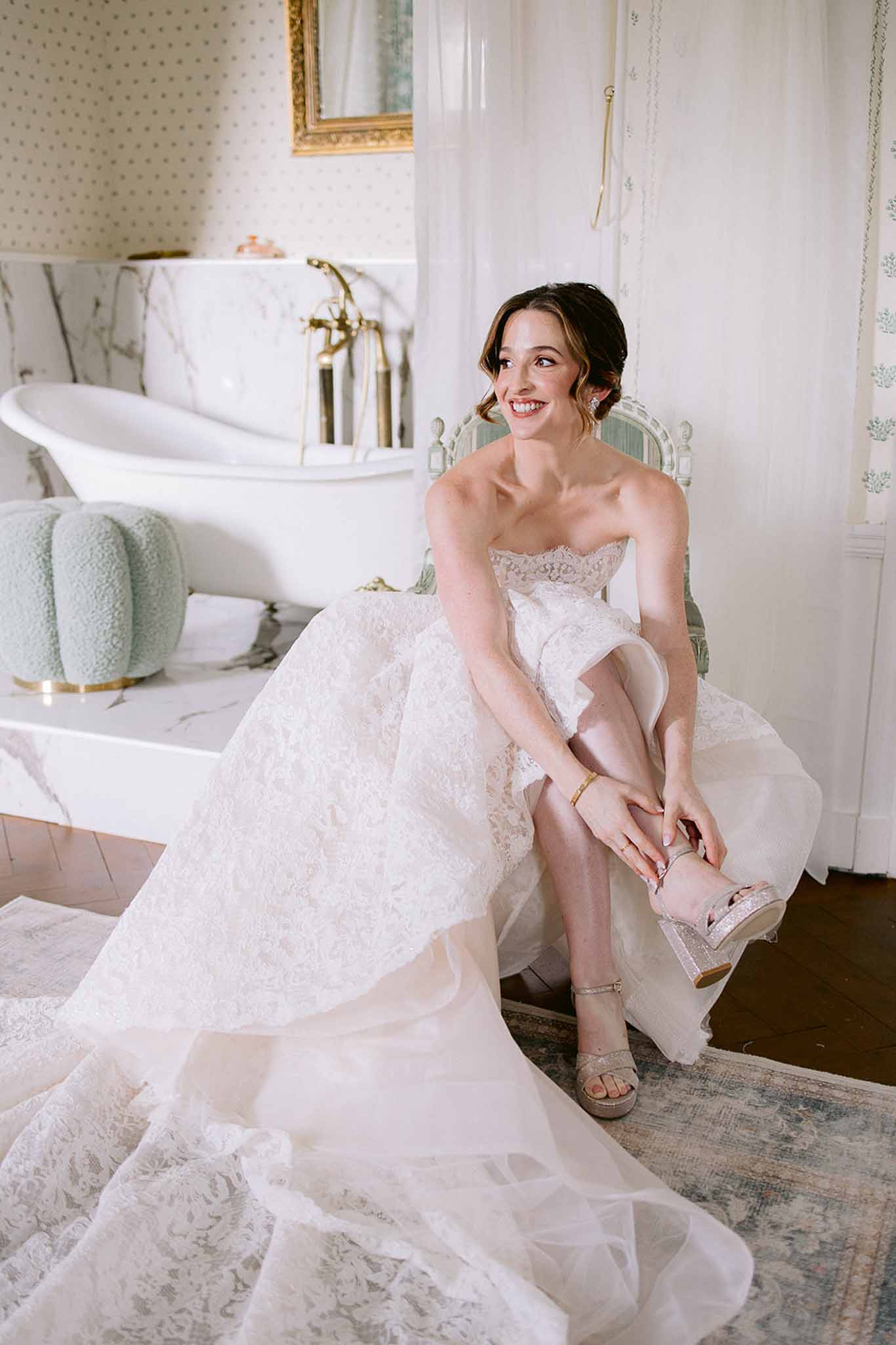 Bride fastening champagne glitter sandal in ivory lace ballgown beside clawfoot tub in polka-dot wallpapered bathroom