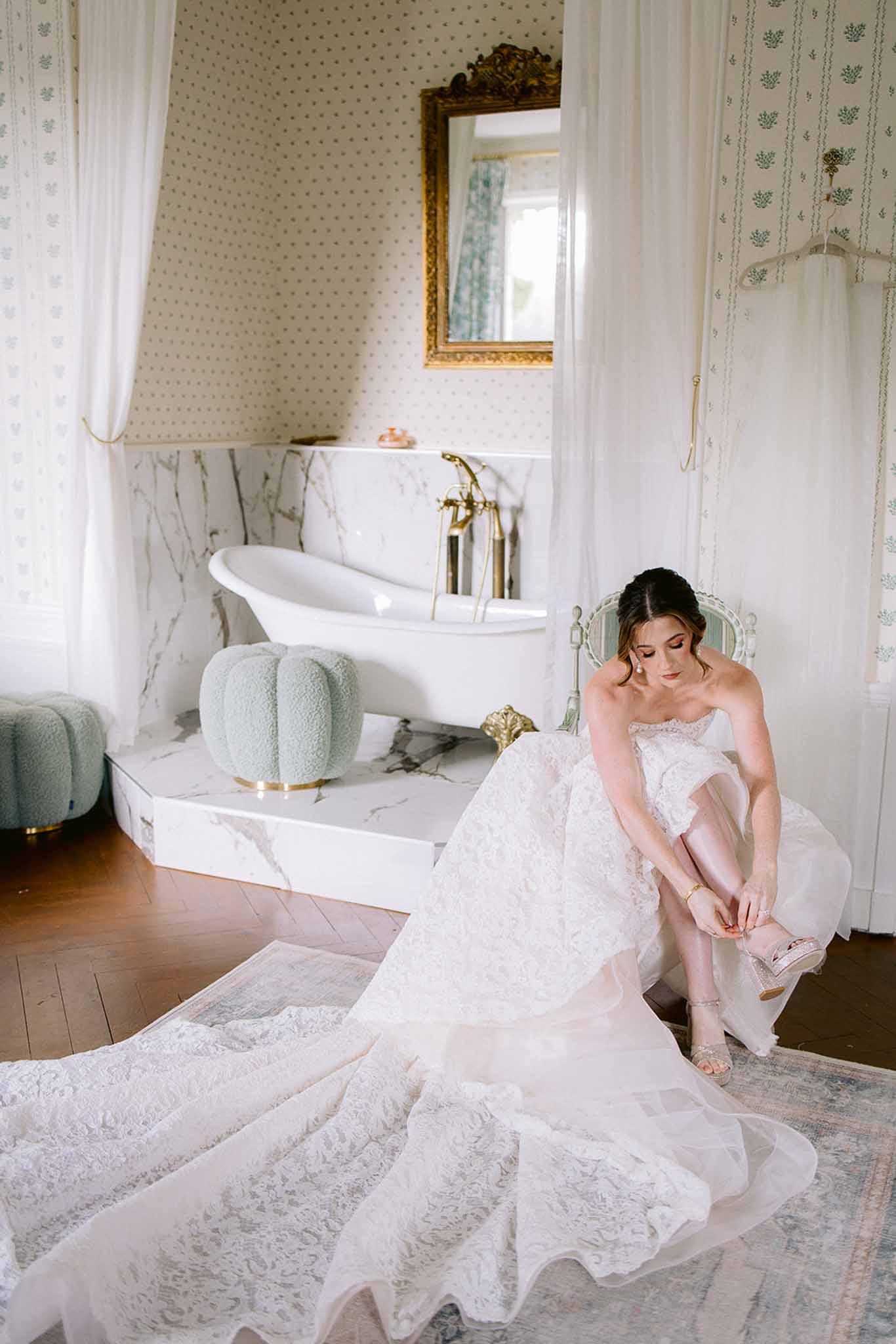 Bride in strapless lace gown fastening sandal in sage green bathroom with clawfoot tub and gold fixtures