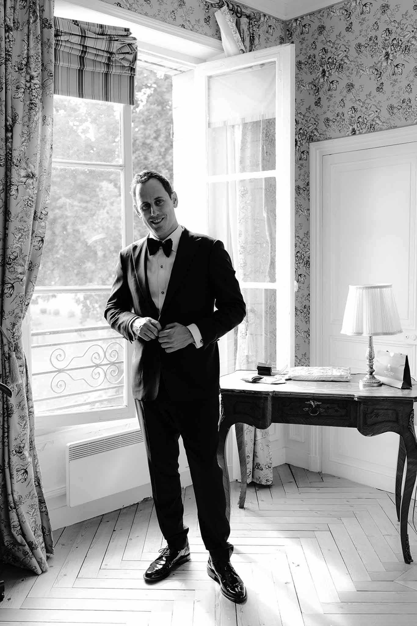 Black and white full-length groom portrait adjusting jacket in floral-wallpapered chateau room