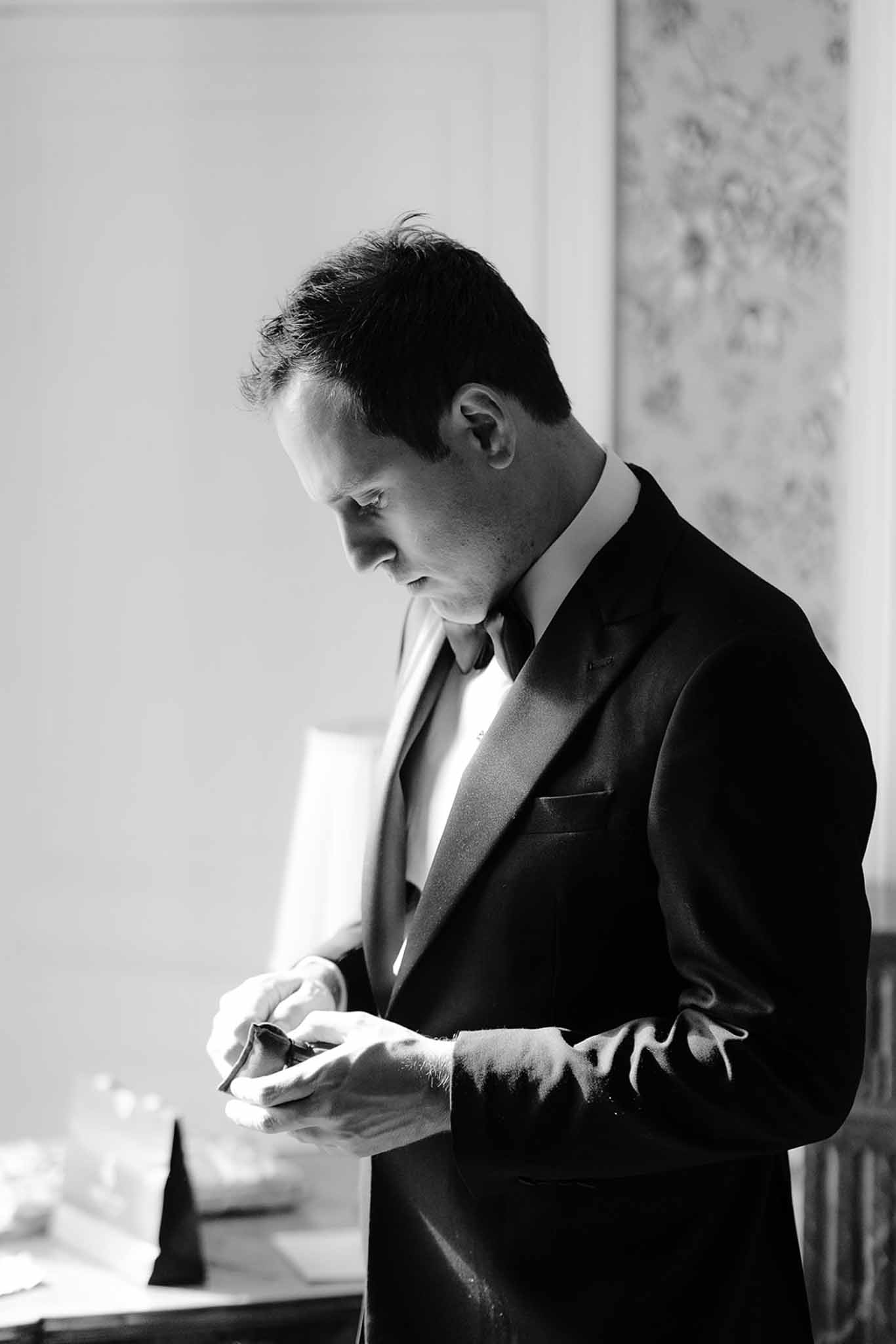 Black and white close-up of groom adjusting cufflinks in dinner jacket by window light