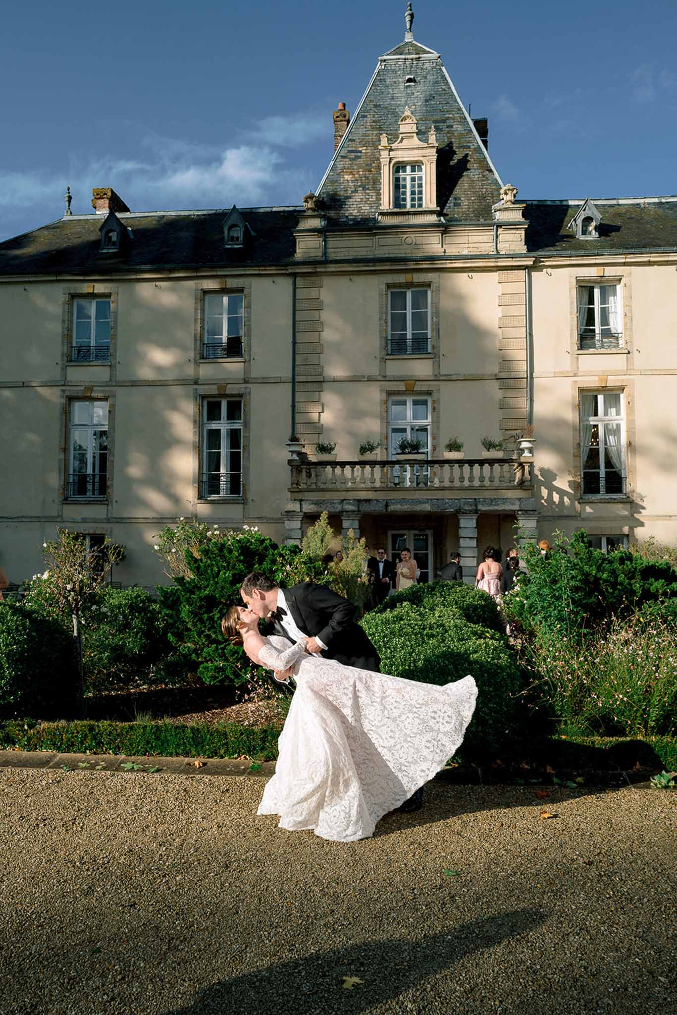 Groom dipping bride into a kiss in formal chateau garden with cream-colored mansion in background