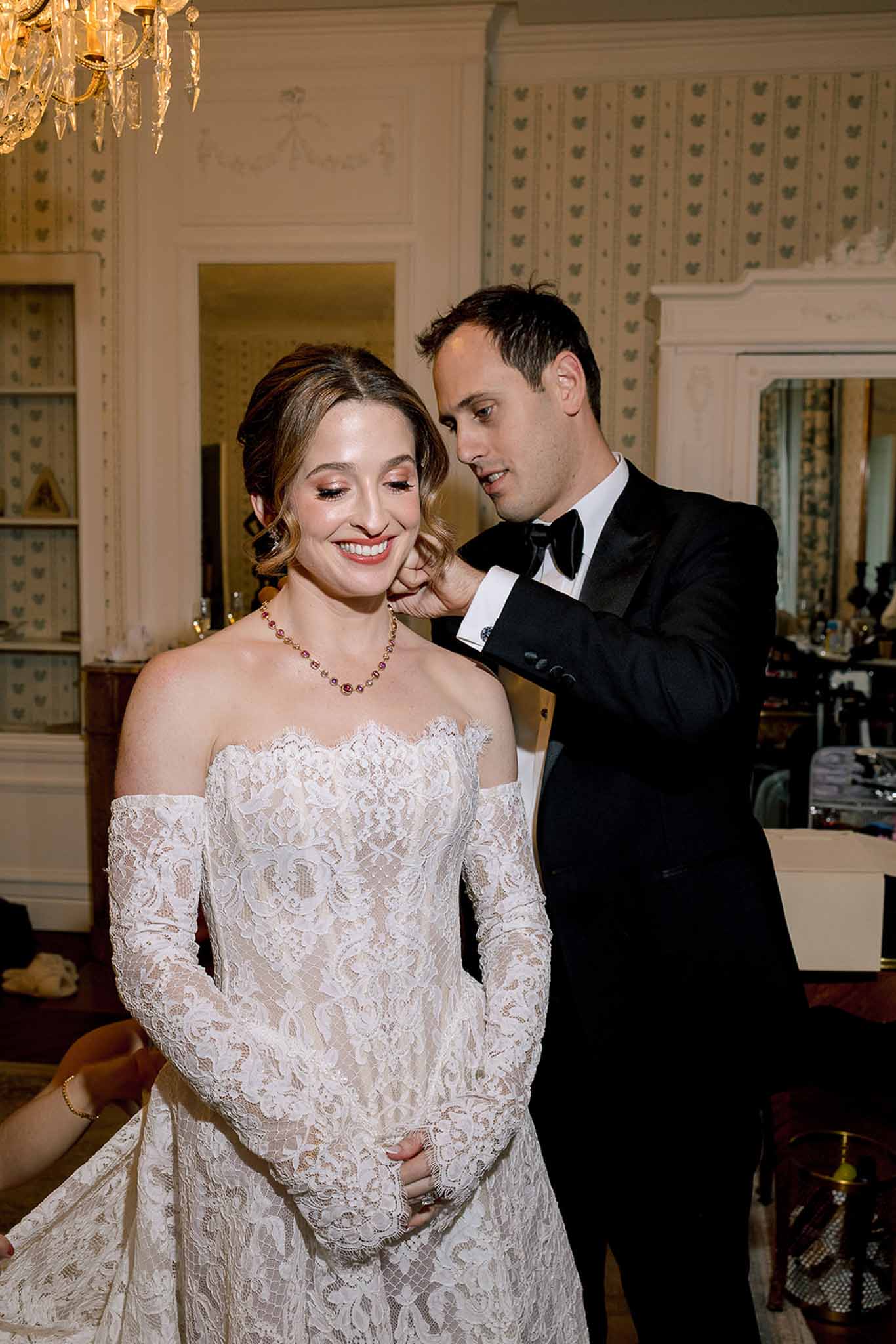 Groom in black tuxedo fastening ruby necklace on smiling bride in off-shoulder lace gown in ornate room