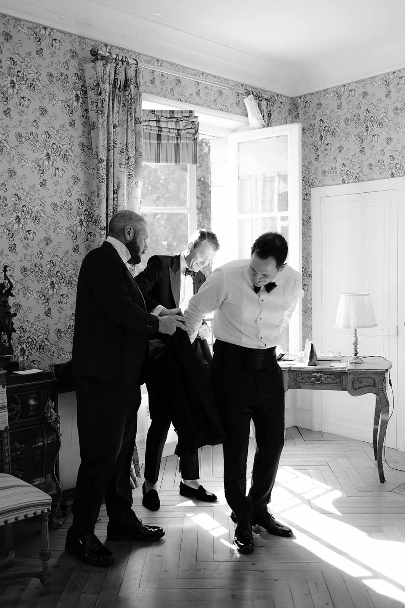 Three men laugh adjusting tuxedos in floral-wallpapered chateau room with parquet floor in B&W