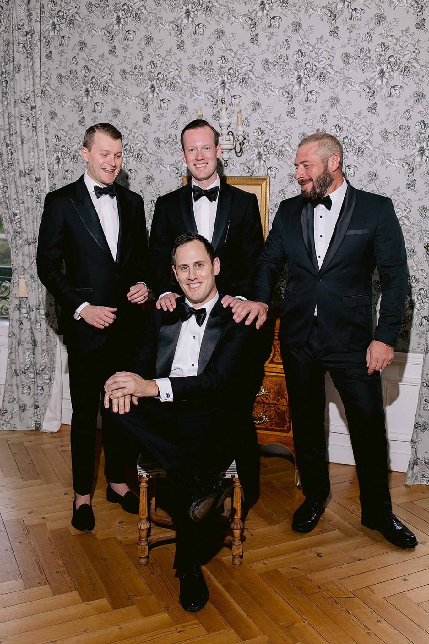 Groom and three groomsmen in black tuxedos posing in chateau room with toile wallpaper