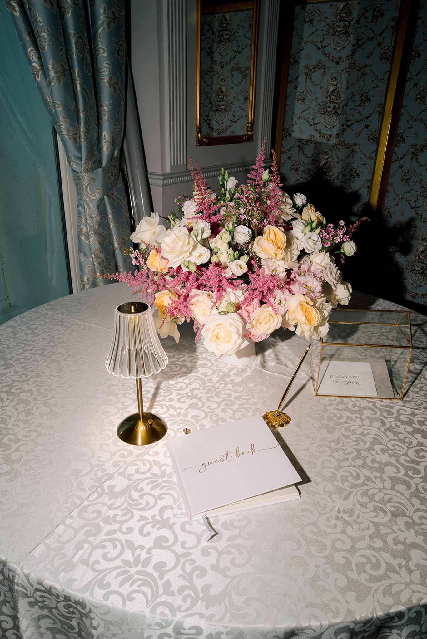 Guest book table with cream and peach garden roses brass lamp and gold script book against blue damask wallpaper