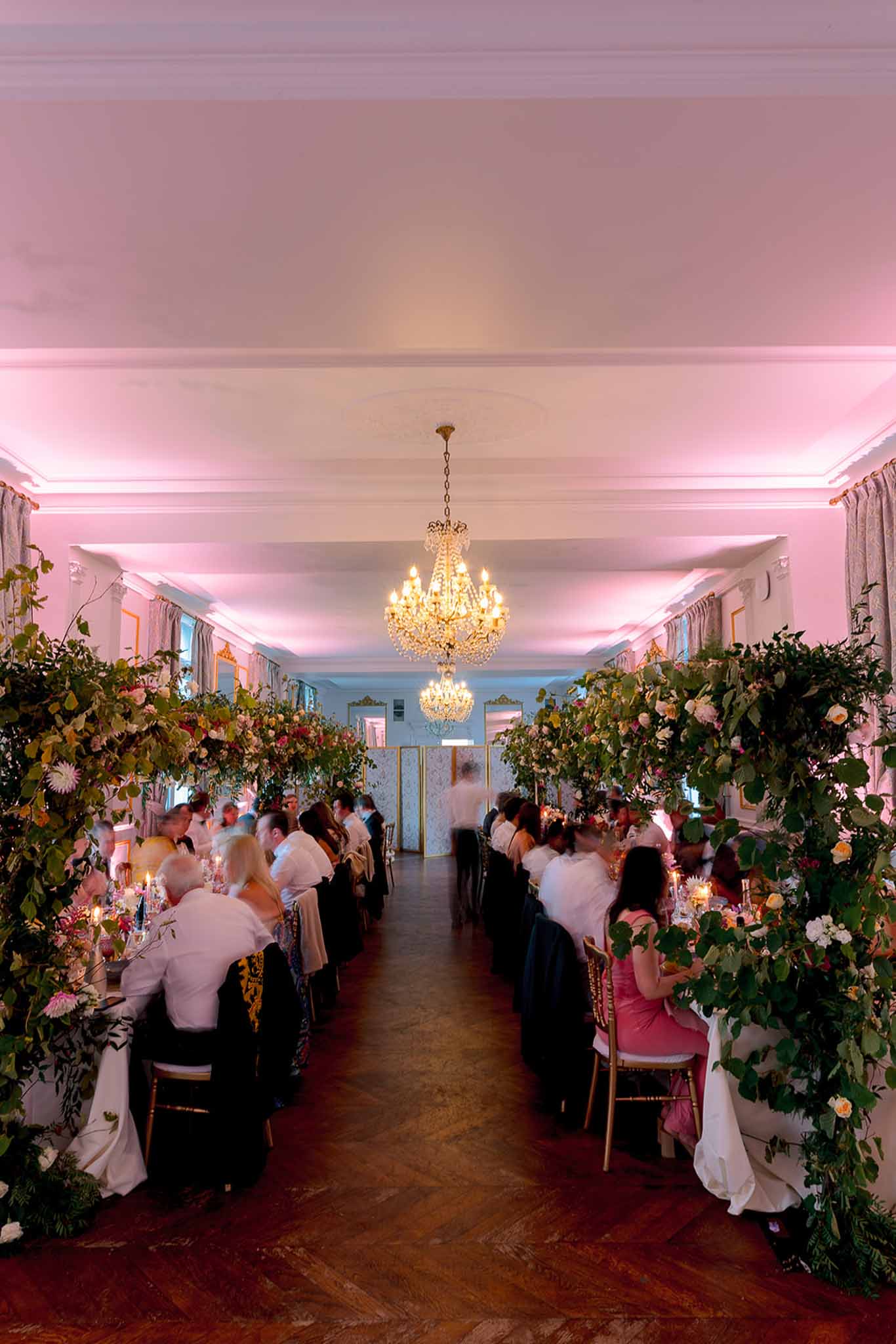 Ballroom reception with long banquet tables under crystal chandeliers and towering peach and blush floral installations