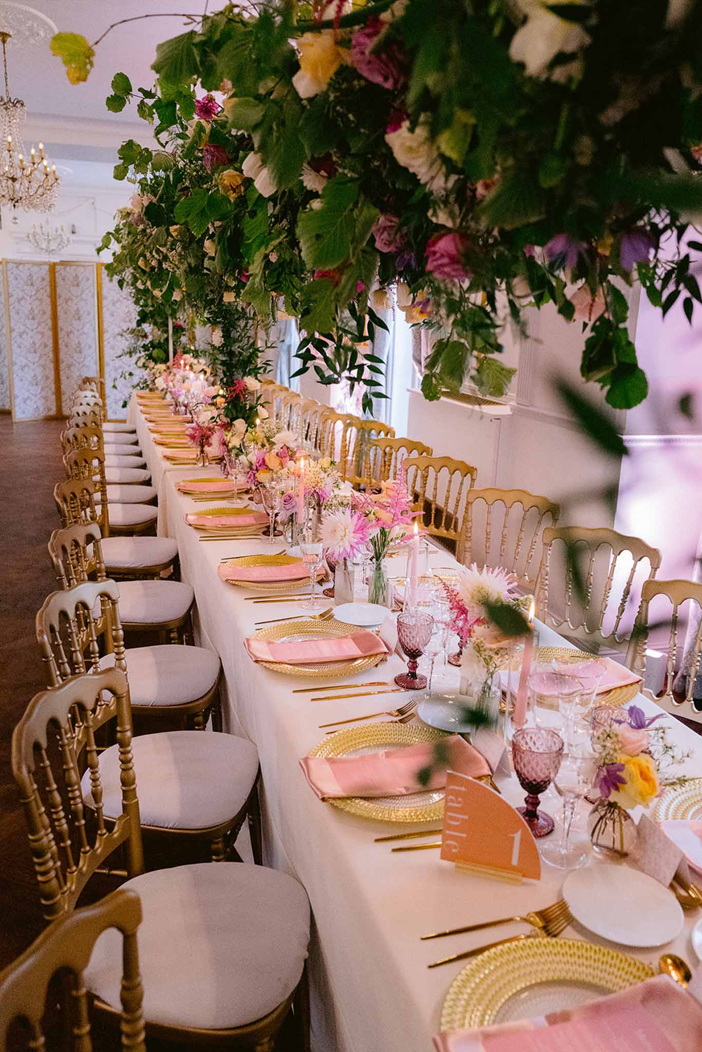 Long banquet table with gold chargers, pink napkins, and suspended floral installation of roses and greenery