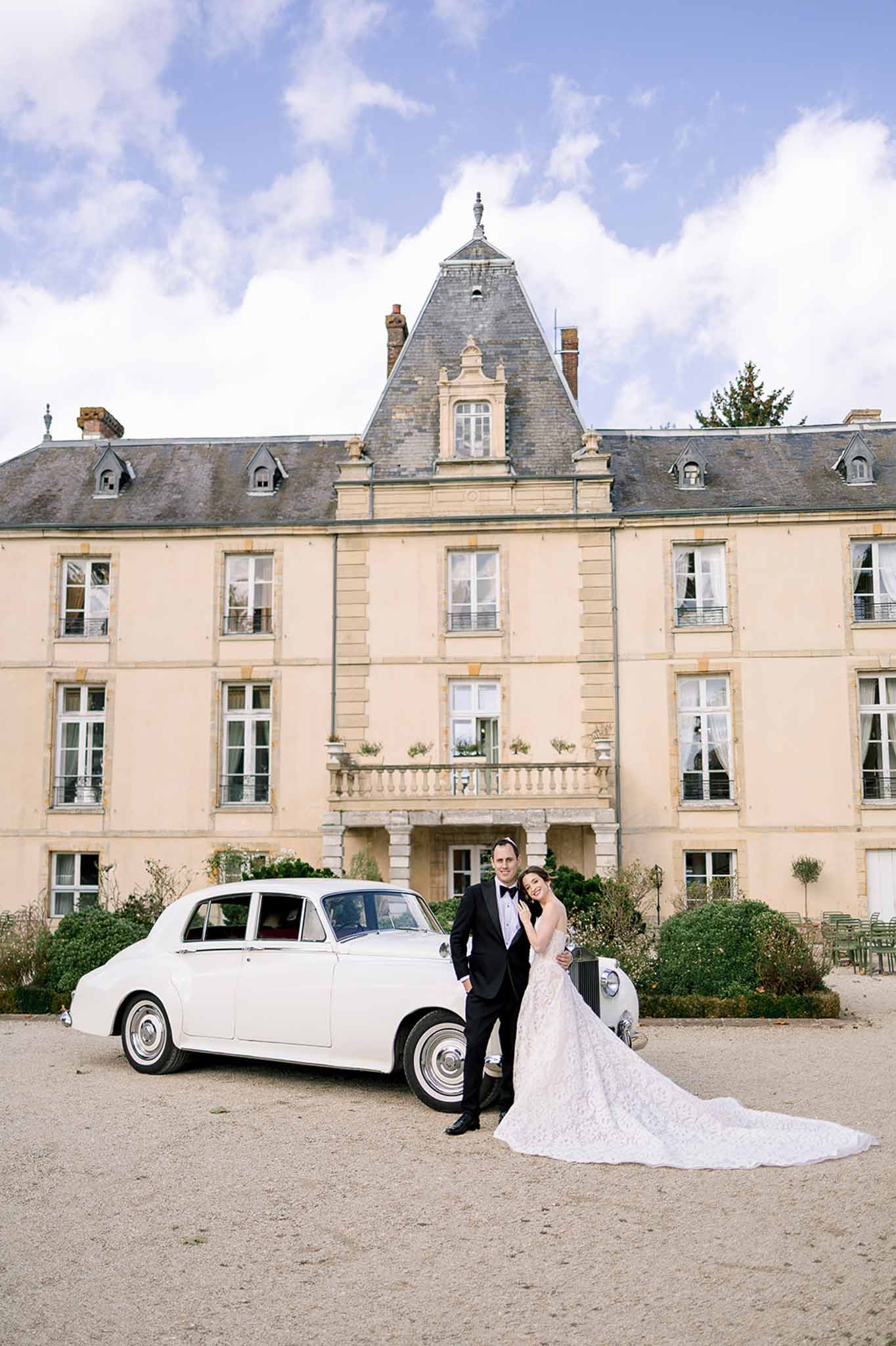 Bride in lace ball gown and groom in tuxedo posing beside a vintage white Rolls-Royce in front of a French chateau