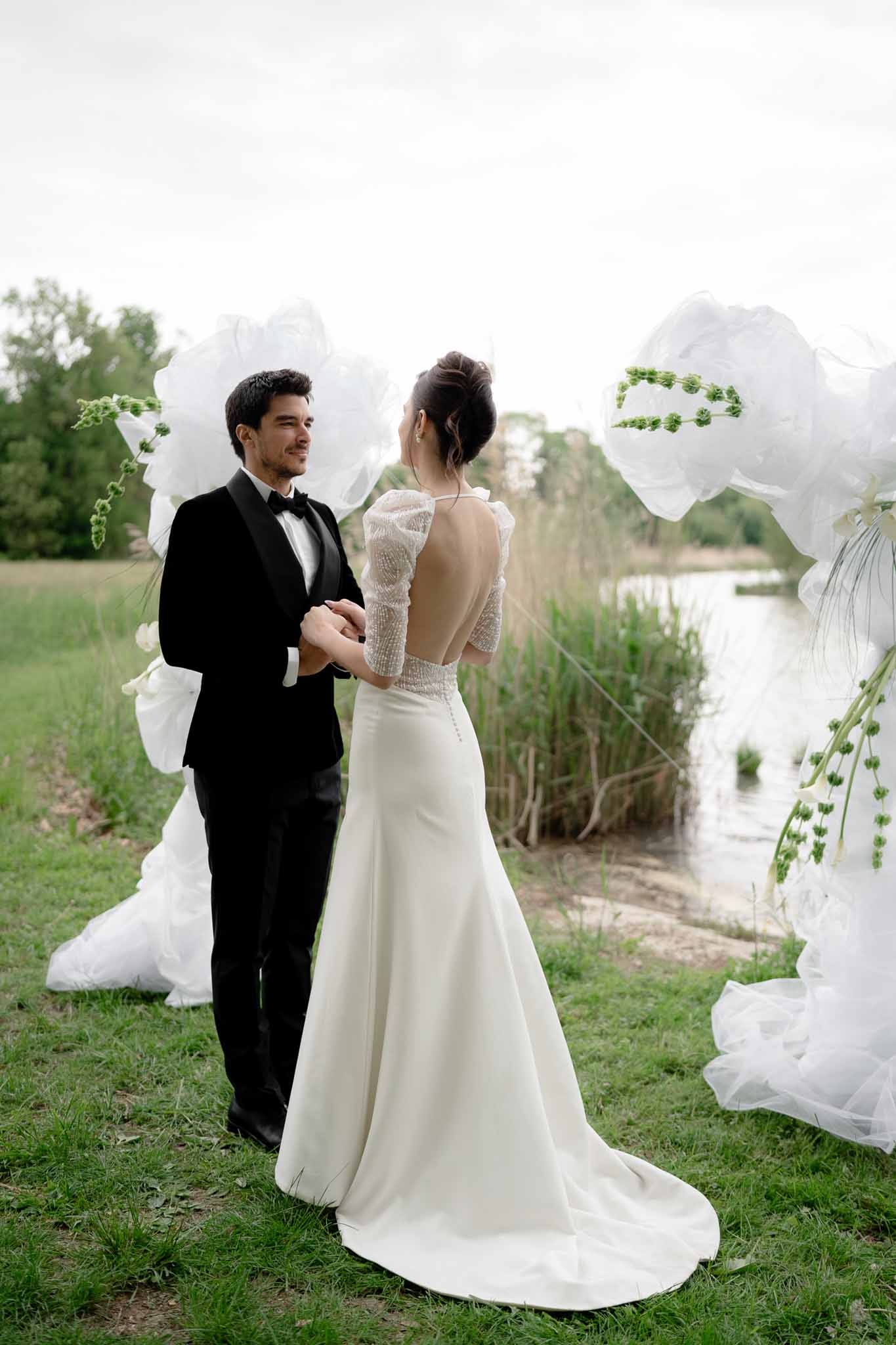Couple holding hands beneath floral arch during lakeside ceremony