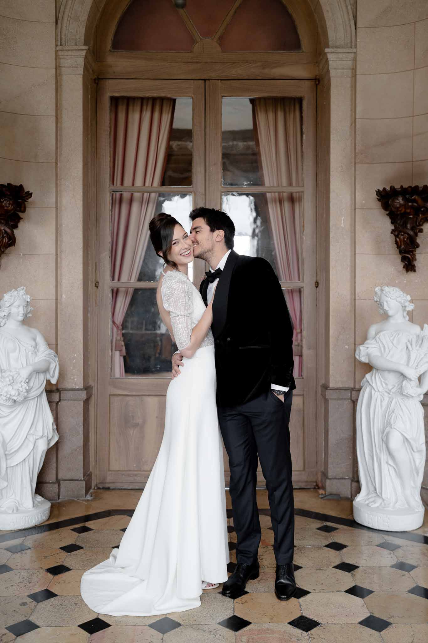 Groom kisses bride's cheek at the grand entrance door of Chateau d'Arcelot