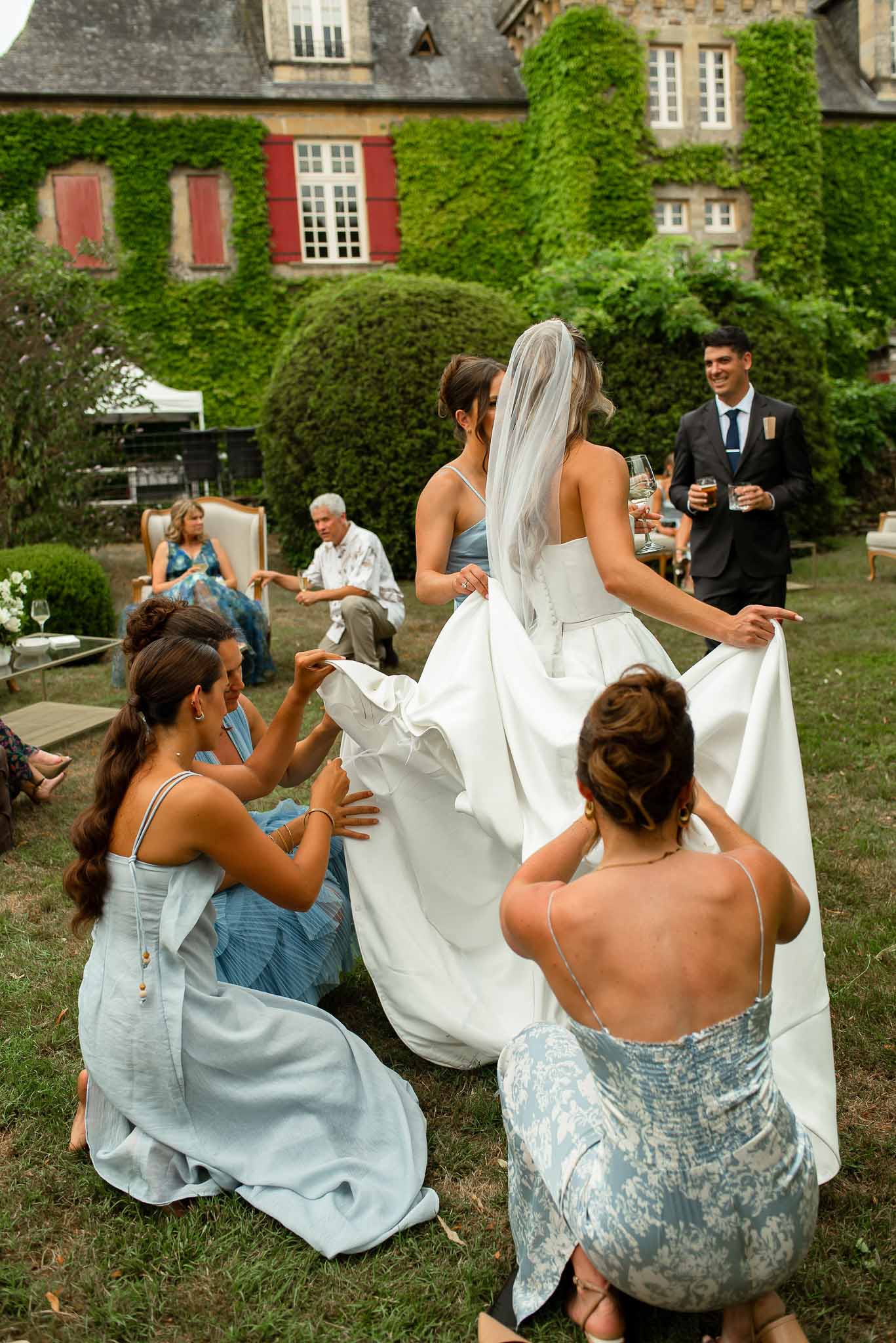 Three bridesmaids in pale blue dresses arranging the bride's white satin gown on a chateau lawn
