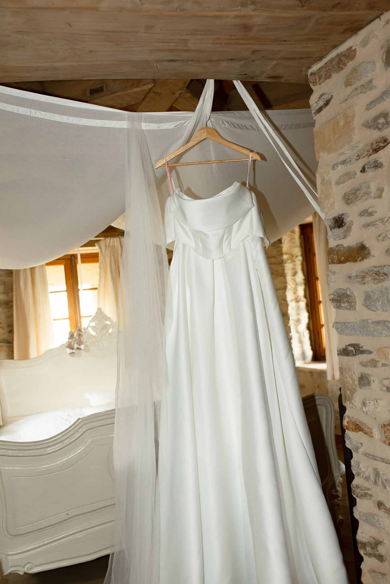 Ivory satin gown with square neckline hanging on four-poster bed in stone-walled rustic bedroom