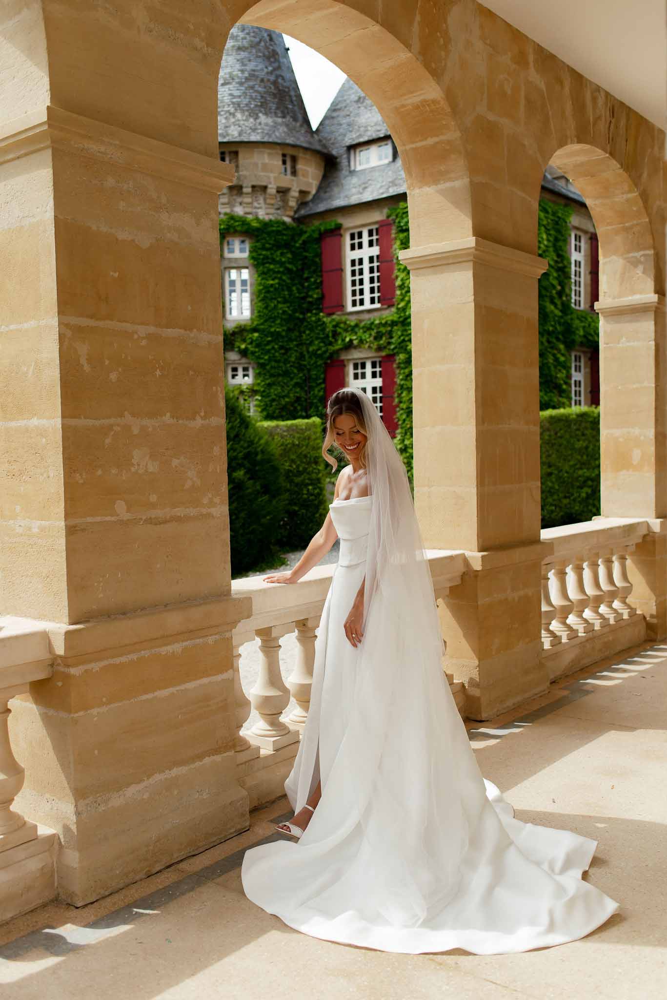 Bride in off-the-shoulder gown with cathedral train on chateau stone terrace framed by arched colonnade