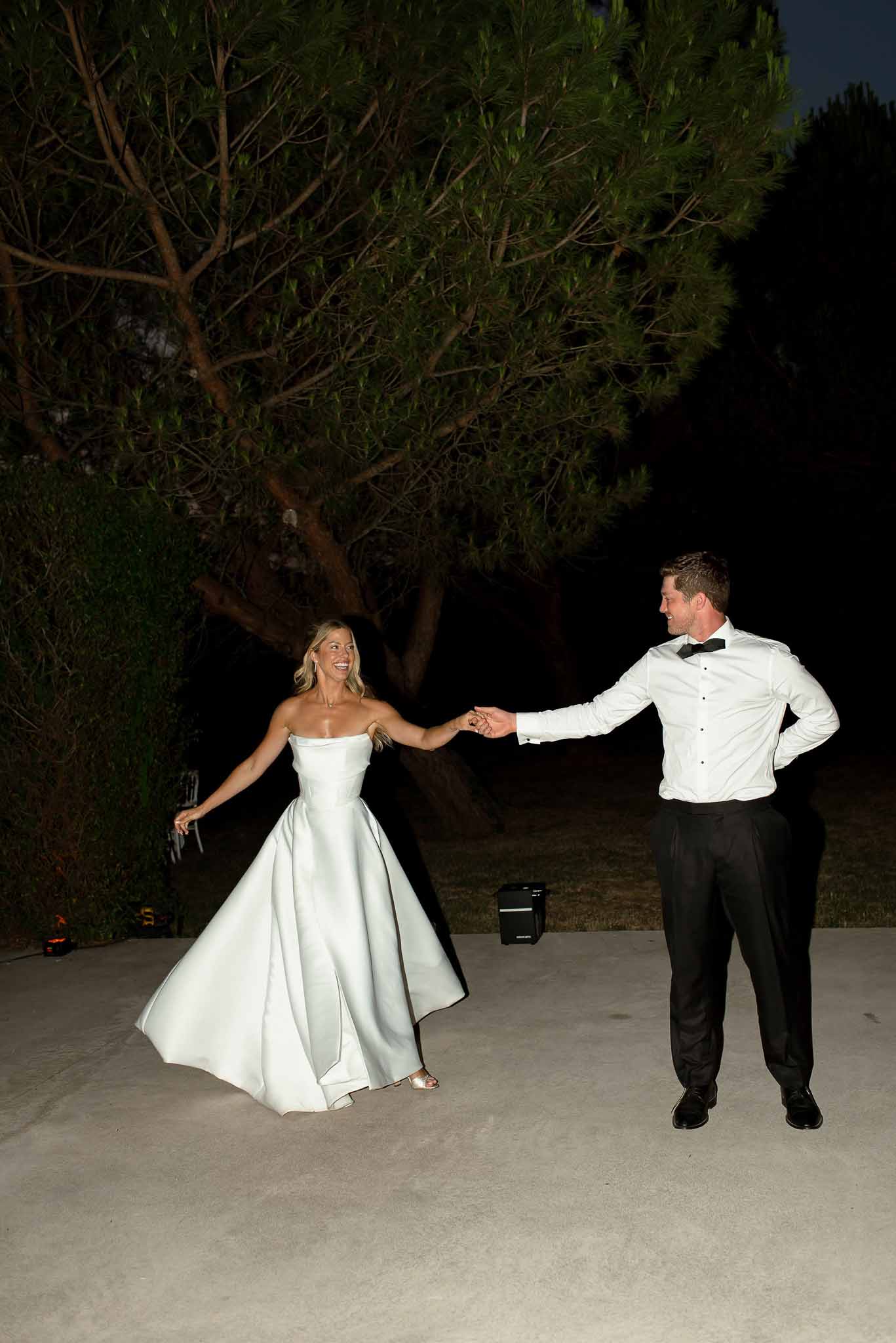 Bride in strapless ballgown twirling with groom on terrace at night under artificial lighting