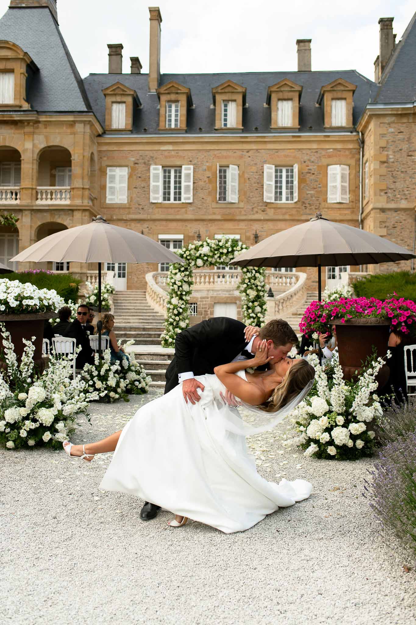 Couple dip kiss after ceremony flanked by white hydrangea arrangements before floral arch and chateau staircase