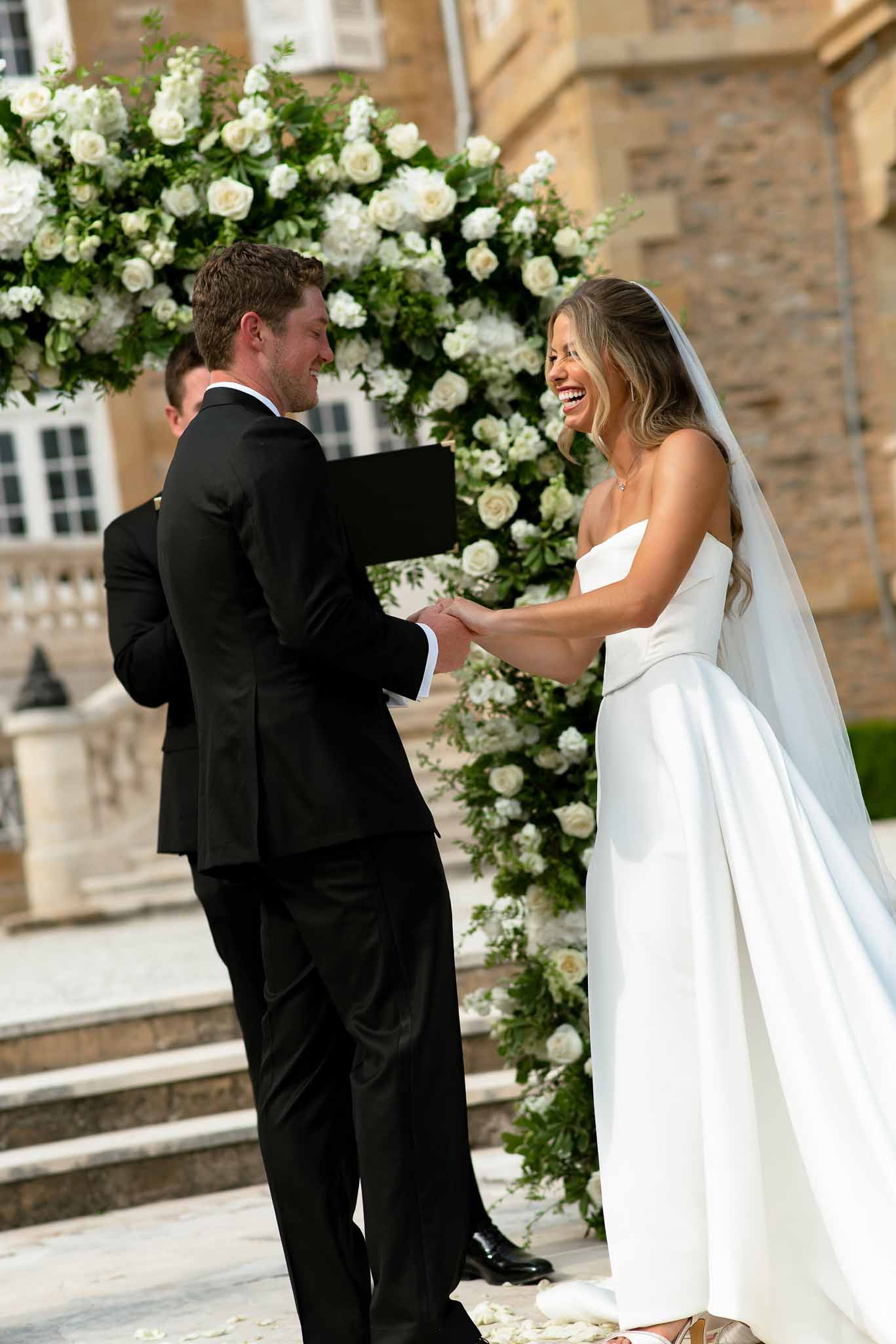 Bride laughing during vow exchange under circular white rose arch at chateau ceremony