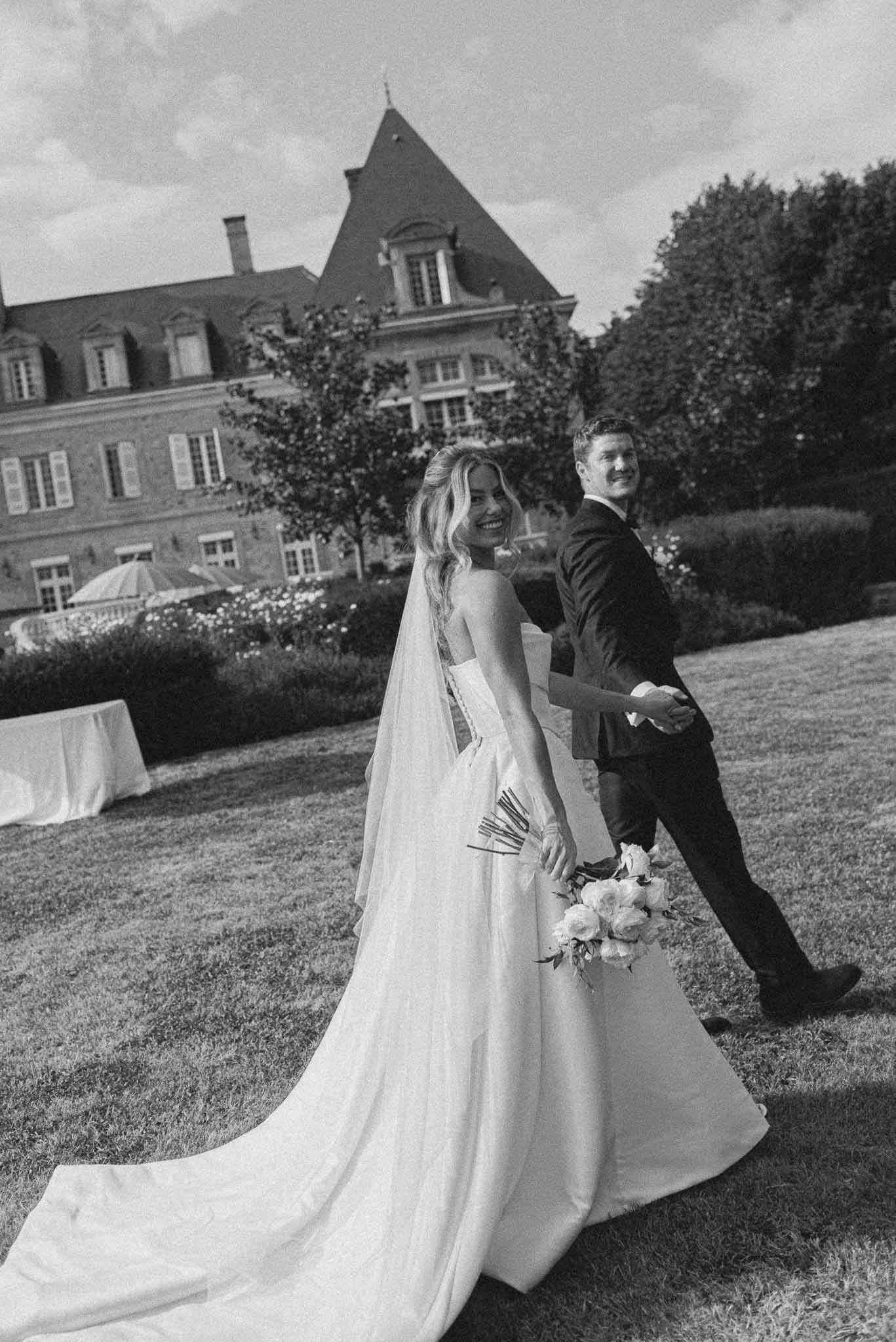 Black-and-white portrait of bride and groom walking hand-in-hand across a chateau lawn, bride turning back and smiling