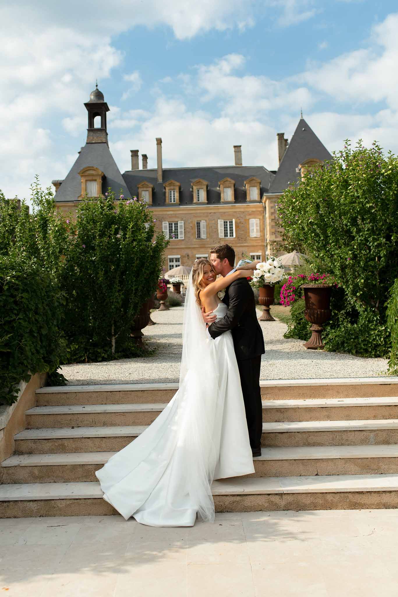 Groom in black suit kisses bride on cheek on chateau terrace steps, bride holds white bouquet with cathedral veil