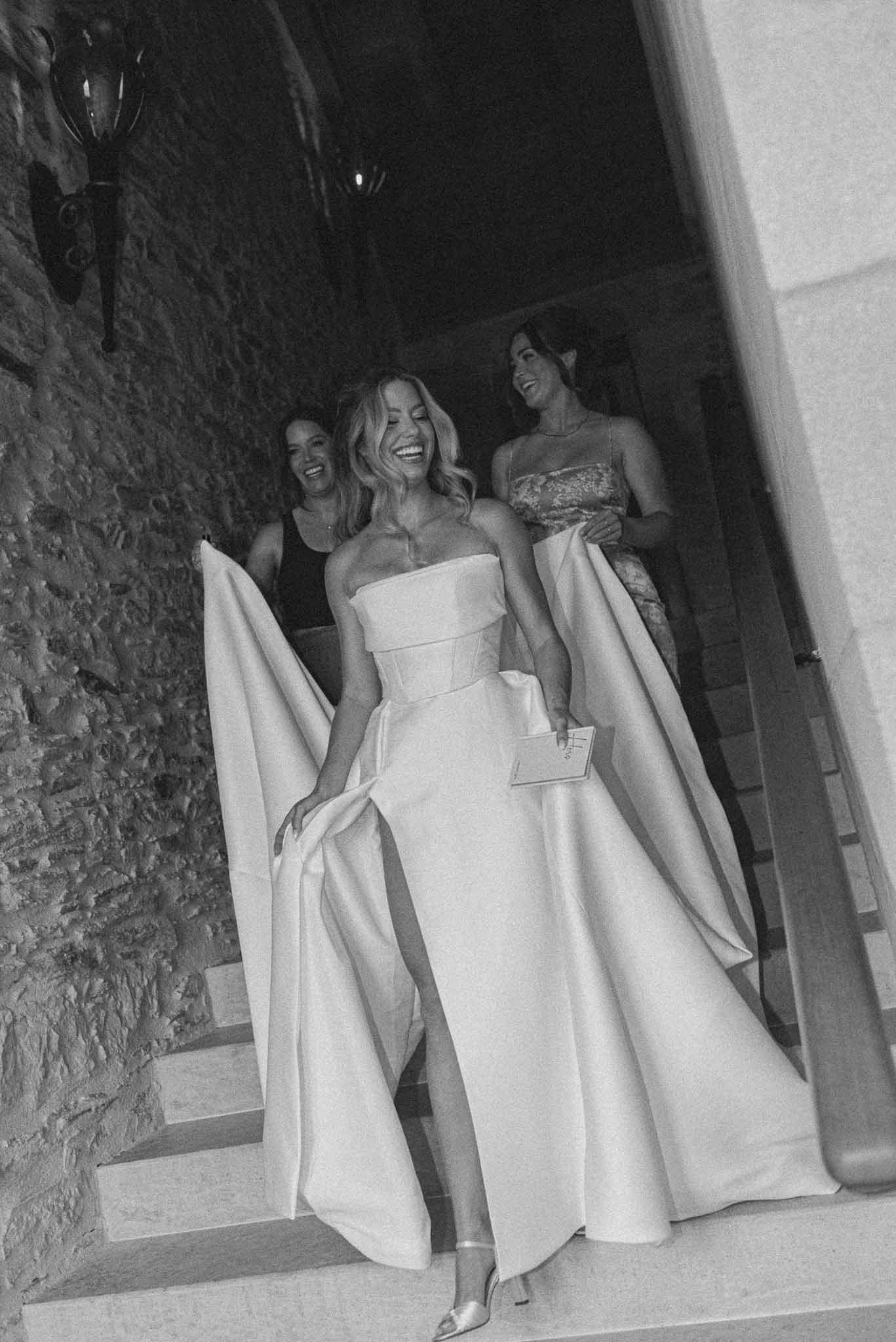 Black and white of bride laughing on stone steps as two women lift her gown train at chateau venue