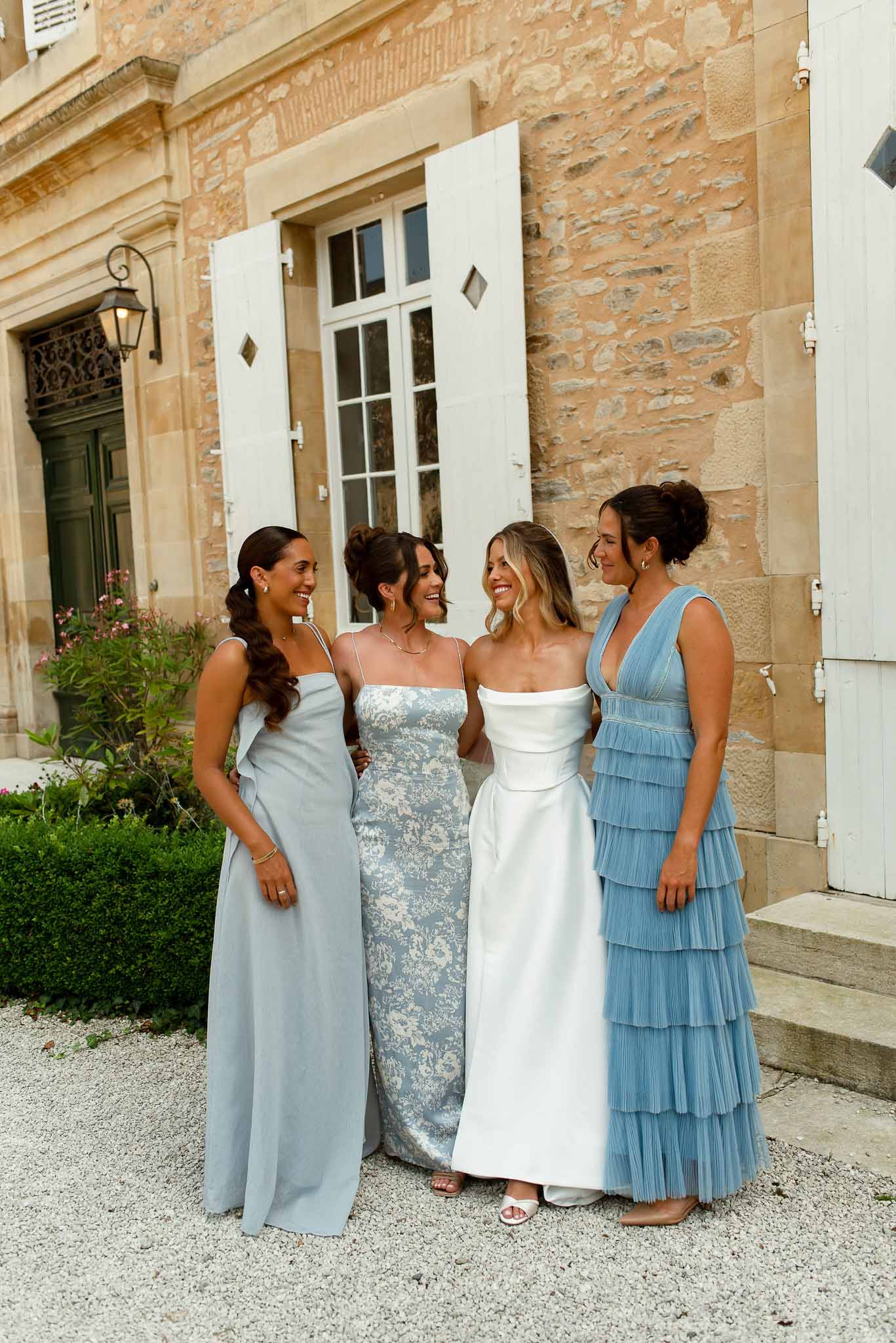 Bride in ivory satin gown with three bridesmaids in mismatched blue dresses before golden chateau