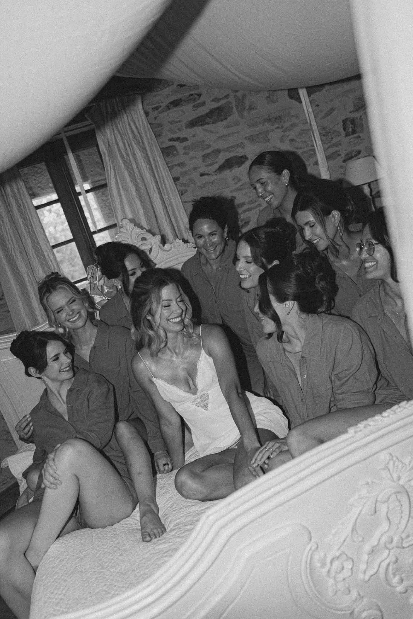 Black and white of bride laughing on ornate bed surrounded by nine bridesmaids in matching shirts
