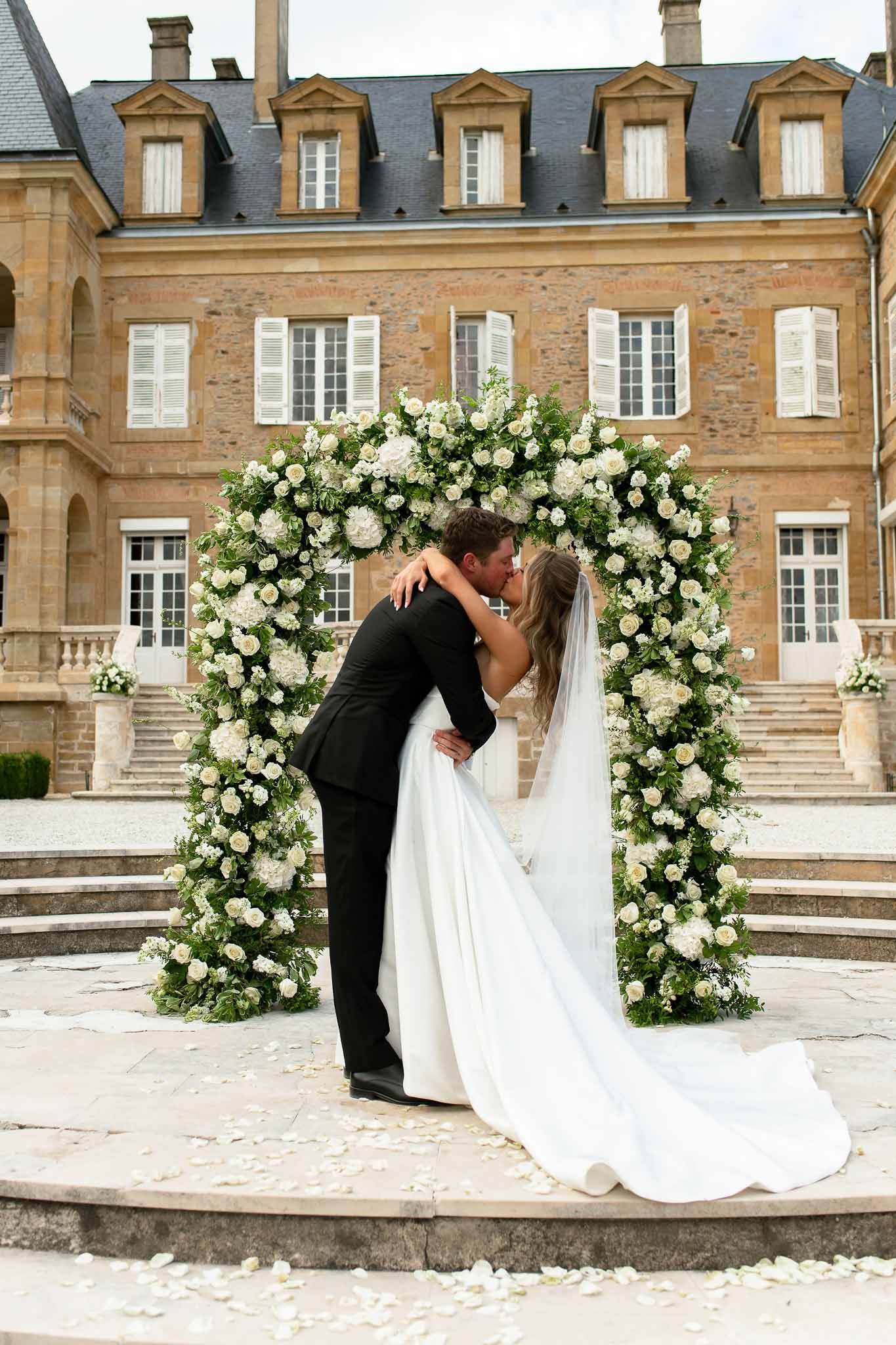 Couple kissing under circular white rose and hydrangea arch with scattered petals before golden stone chateau facade