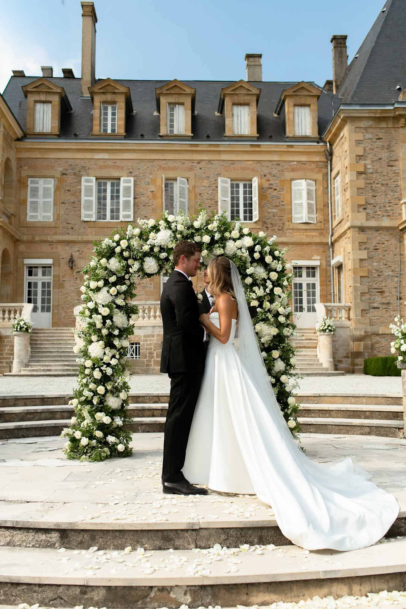 Couple under circular white hydrangea and rose arch before golden limestone chateau with grand staircase