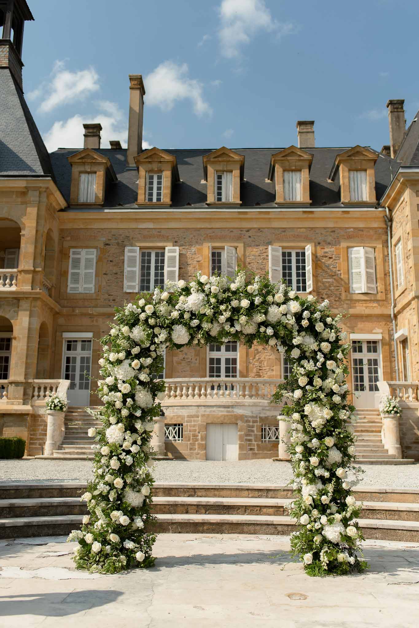 Circular floral arch of white roses and hydrangeas set before golden limestone chateau facade with stone staircases
