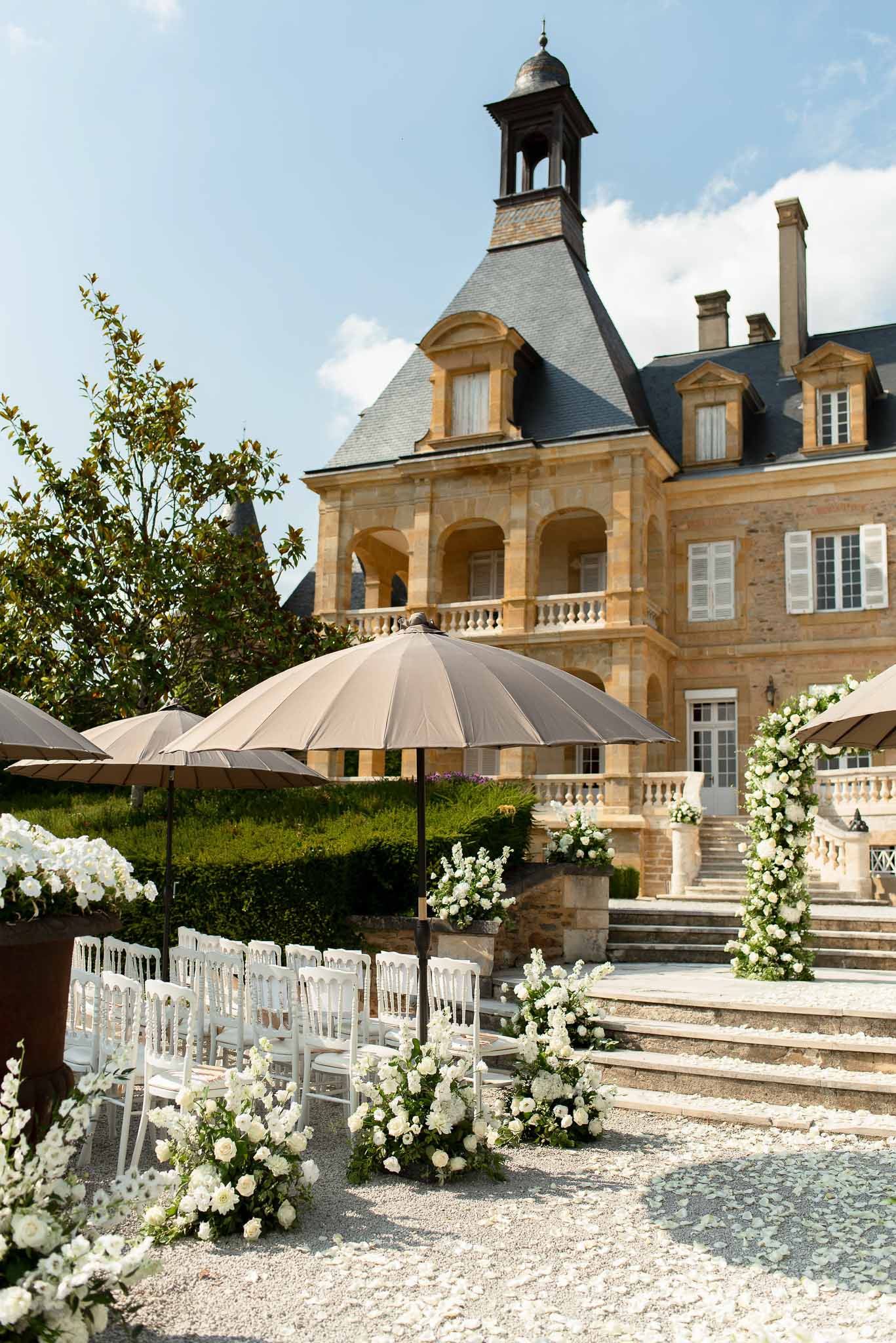 White Napoleon chairs with petal aisle and floral arch on gravel before golden limestone chateau
