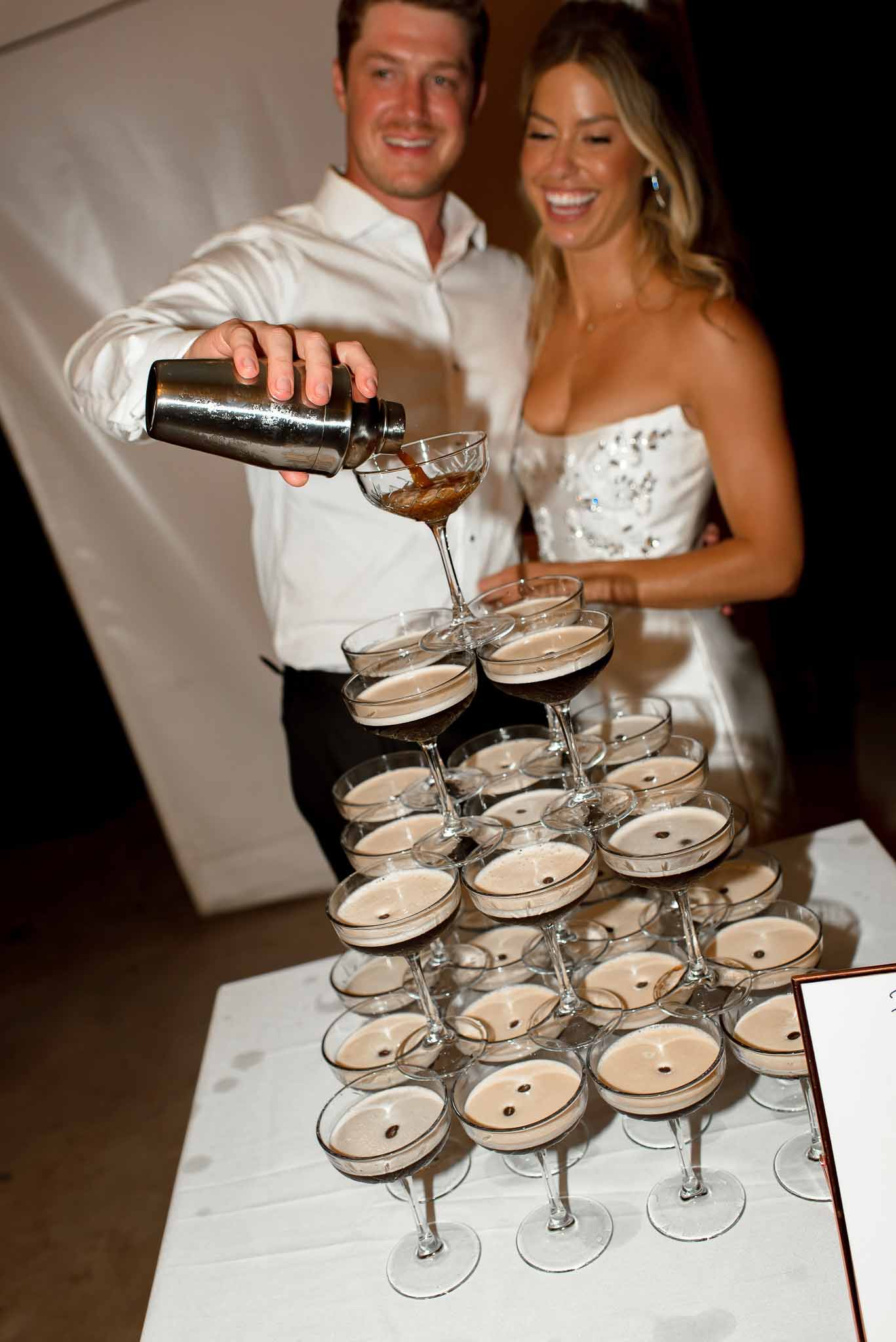 Bride and groom pouring espresso martini tower of coupe glasses during wedding reception