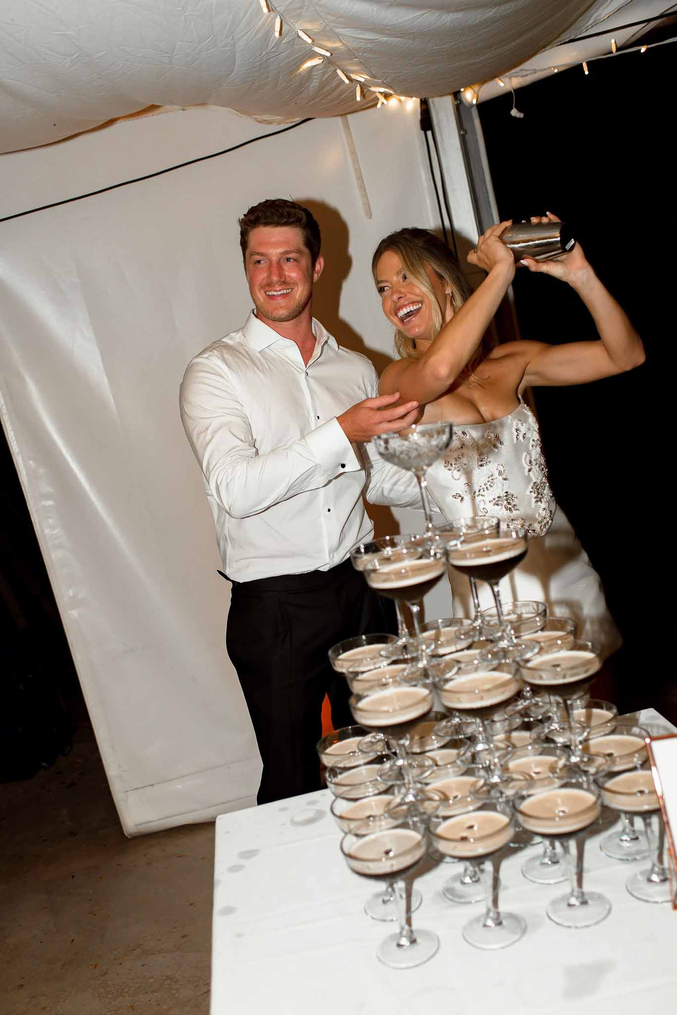 Bride and groom laughing while pouring cocktails into a coupe glass tower inside a marquee with fairy lights