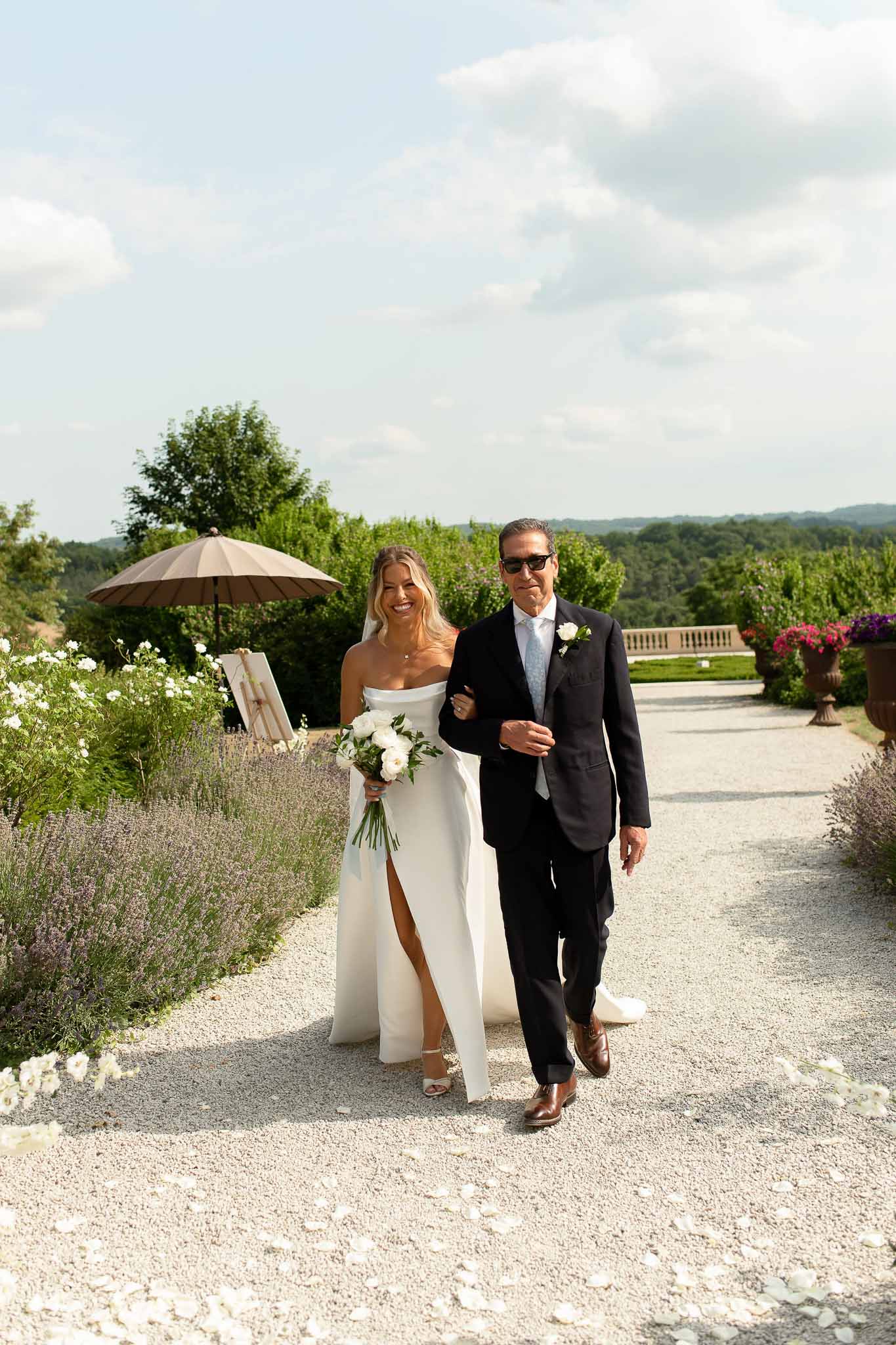 Father walking bride down petal-strewn garden path with white peony bouquet past lavender borders